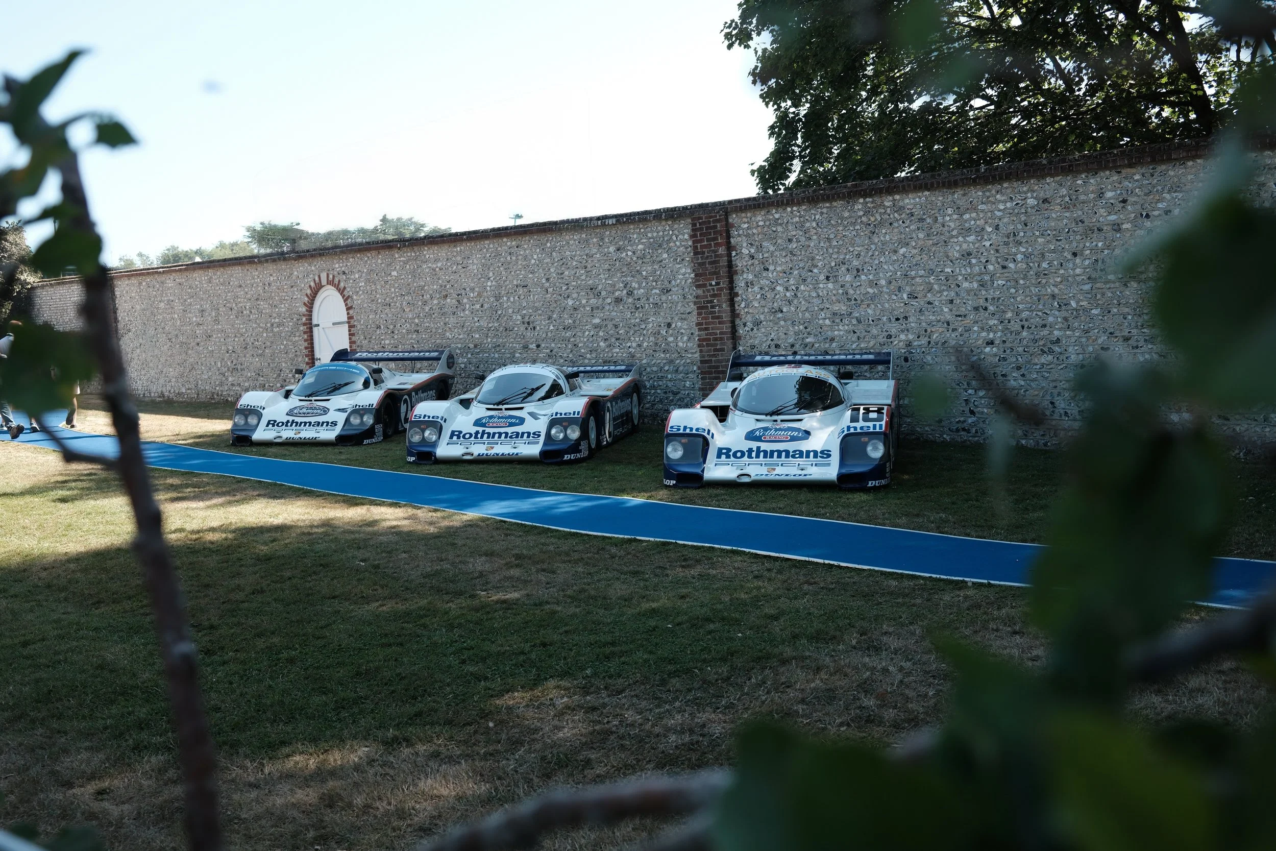 Three vintage race cars with Rothmans logos parked against a stone wall, framed by trees and branches.