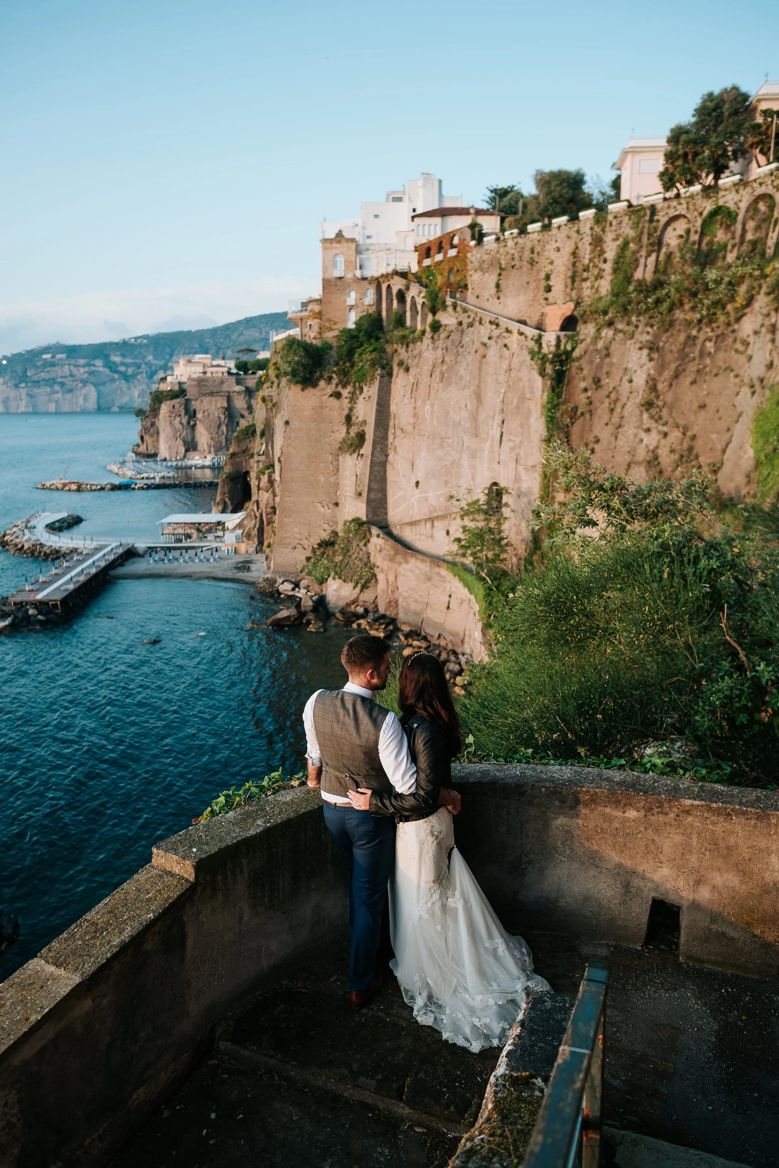 A couple embracing on a stone balcony overlooking a cliffside with buildings and the sea in the background at a wedding at Villa Anticha Mura in Sorrento, Amalfi Coast, Italy