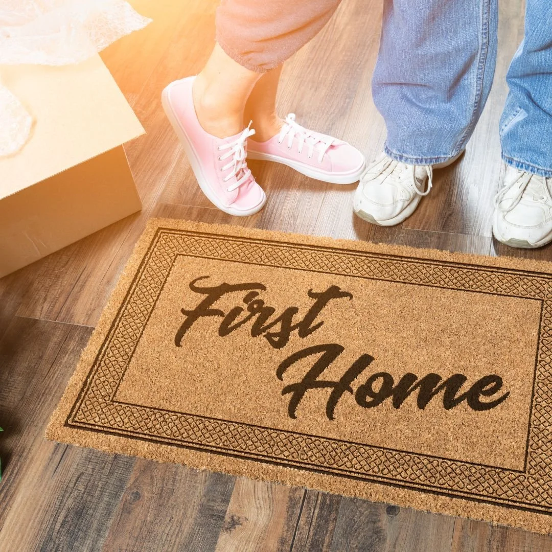 Doormat with text 'first home', pair of feet in background with moving box