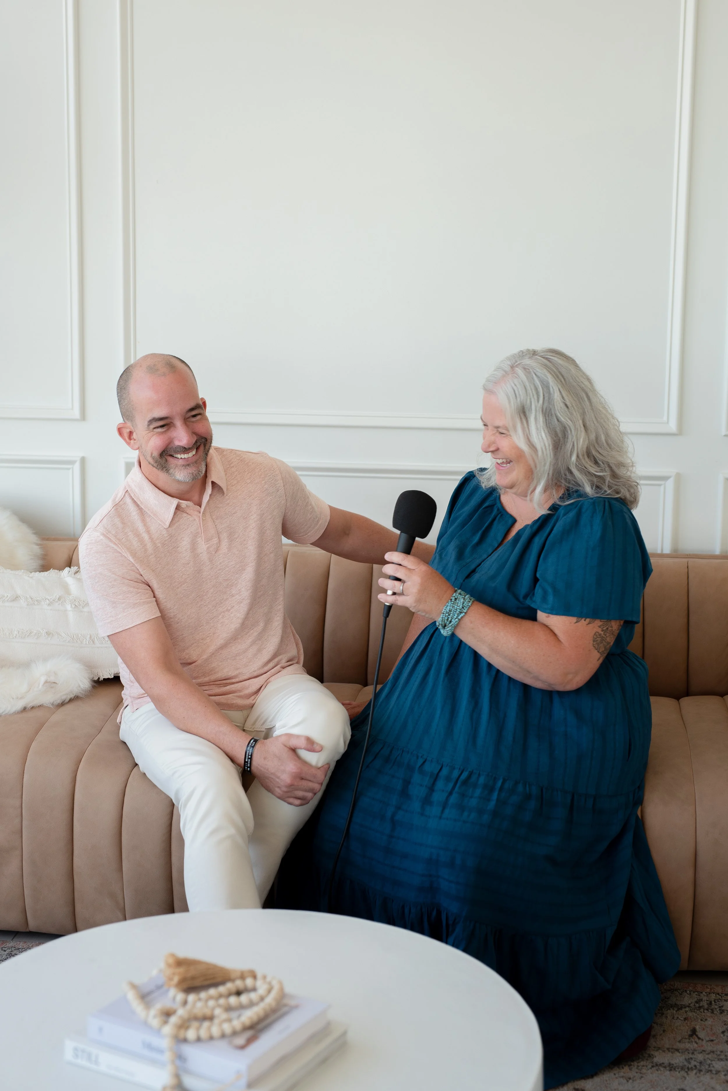 A woman with gray hair in a blue dress is holding a microphone and talking to a smiling man with a shaved head wearing a light pink shirt, sitting on a beige sofa in a bright room with white walls.