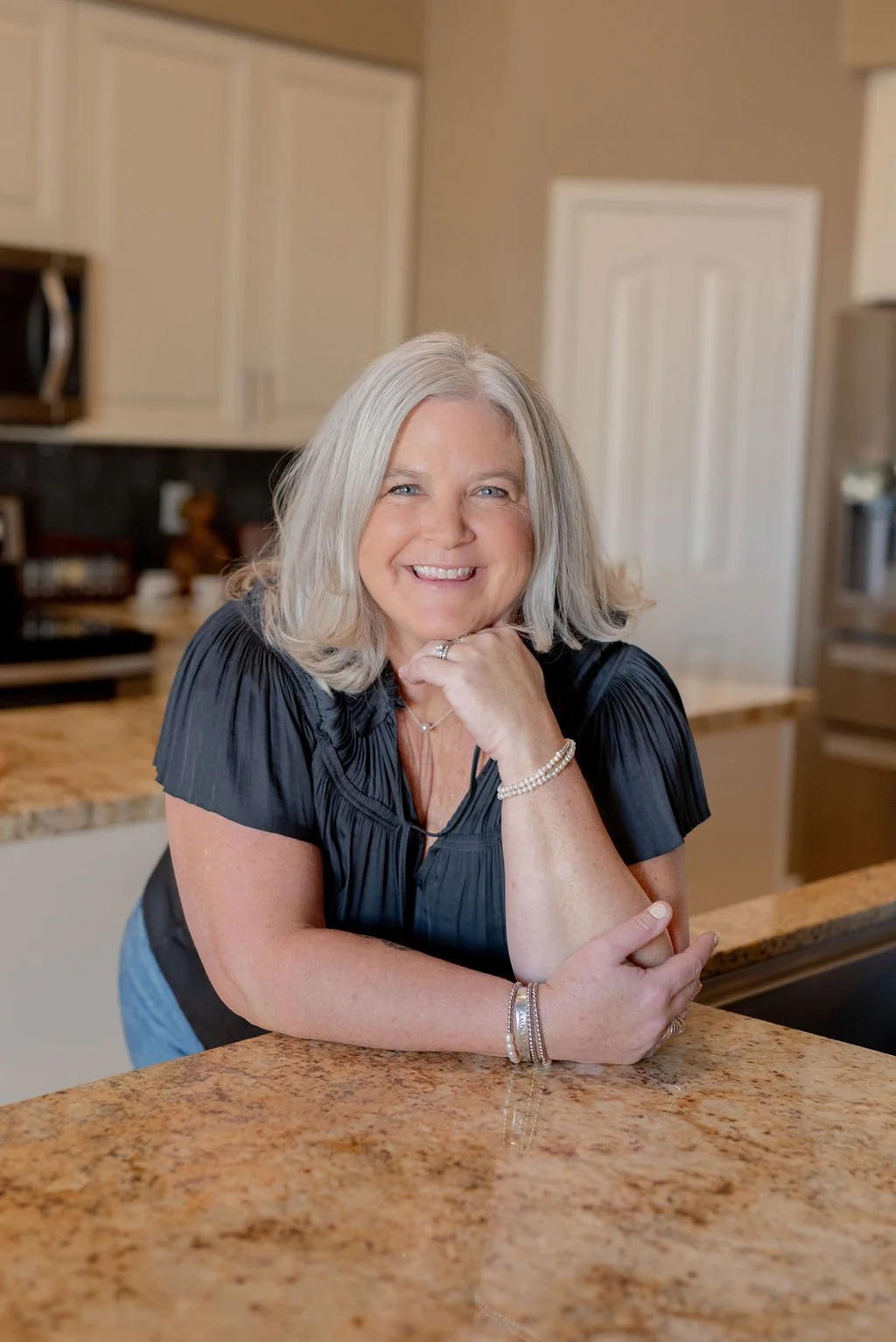 A smiling middle-aged woman with gray hair, wearing a black top and jewelry, leaning on a kitchen counter.