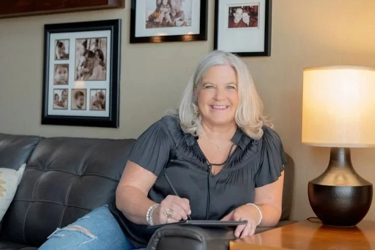 A woman with shoulder-length gray hair sitting on a dark leather couch, smiling, with a lamp and family photos on the wall behind her.