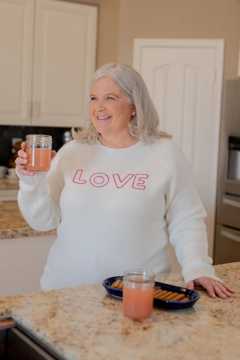 A smiling woman with gray hair in a white sweater with the word 'LOVE' in pink on the front, holding a glass jar with pink beverage, standing in a kitchen with a bowl of cookies and another jar of pink drink on the counter.