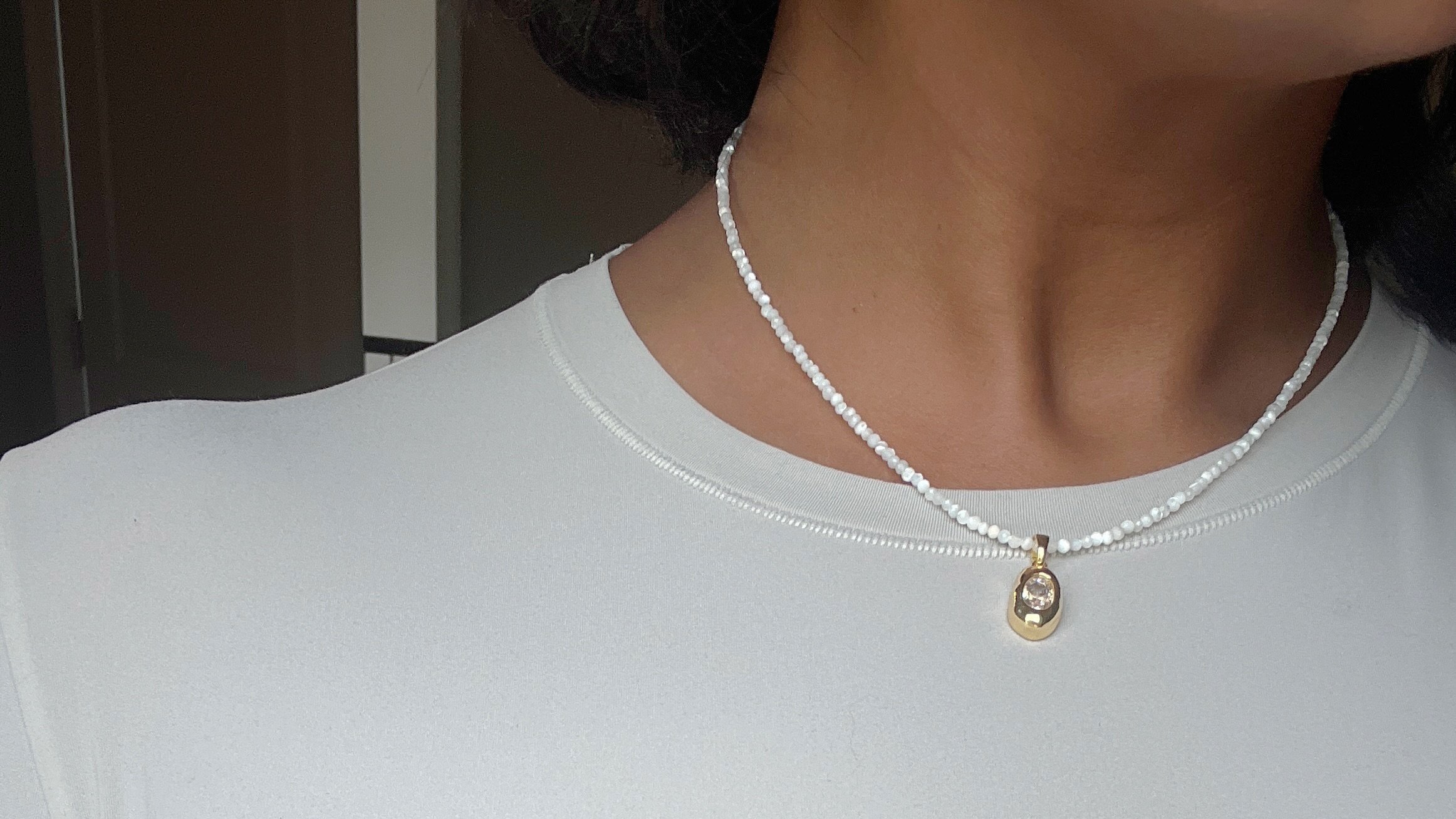 A close up image of a necklace on a woman's neck. The necklace is beaded with small white pearl beads and has an irregular charm on it. The charm is gold and has a CZ stone.