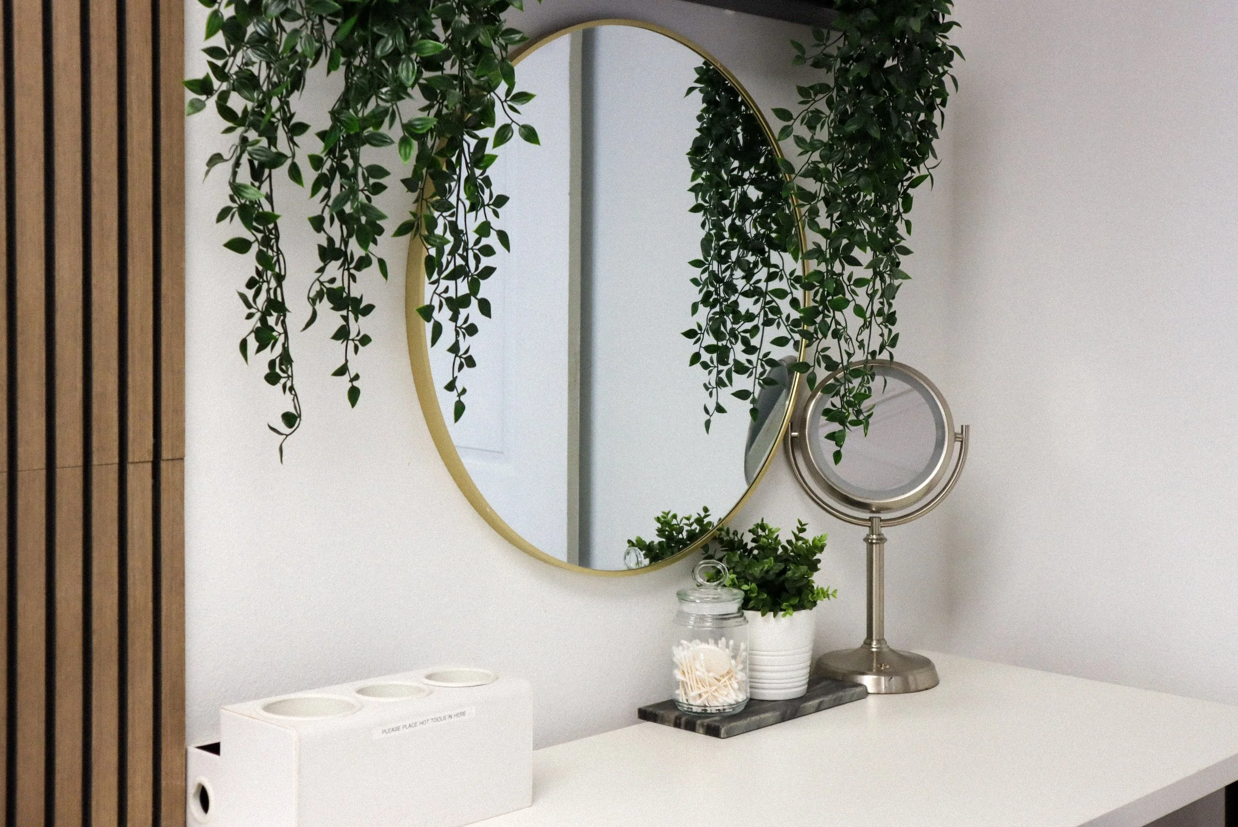 A mirror with multiple round lights around the frame, placed on a white table with dried flowers and jars, in front of a window. Professional lighting equipment is visible in the background.