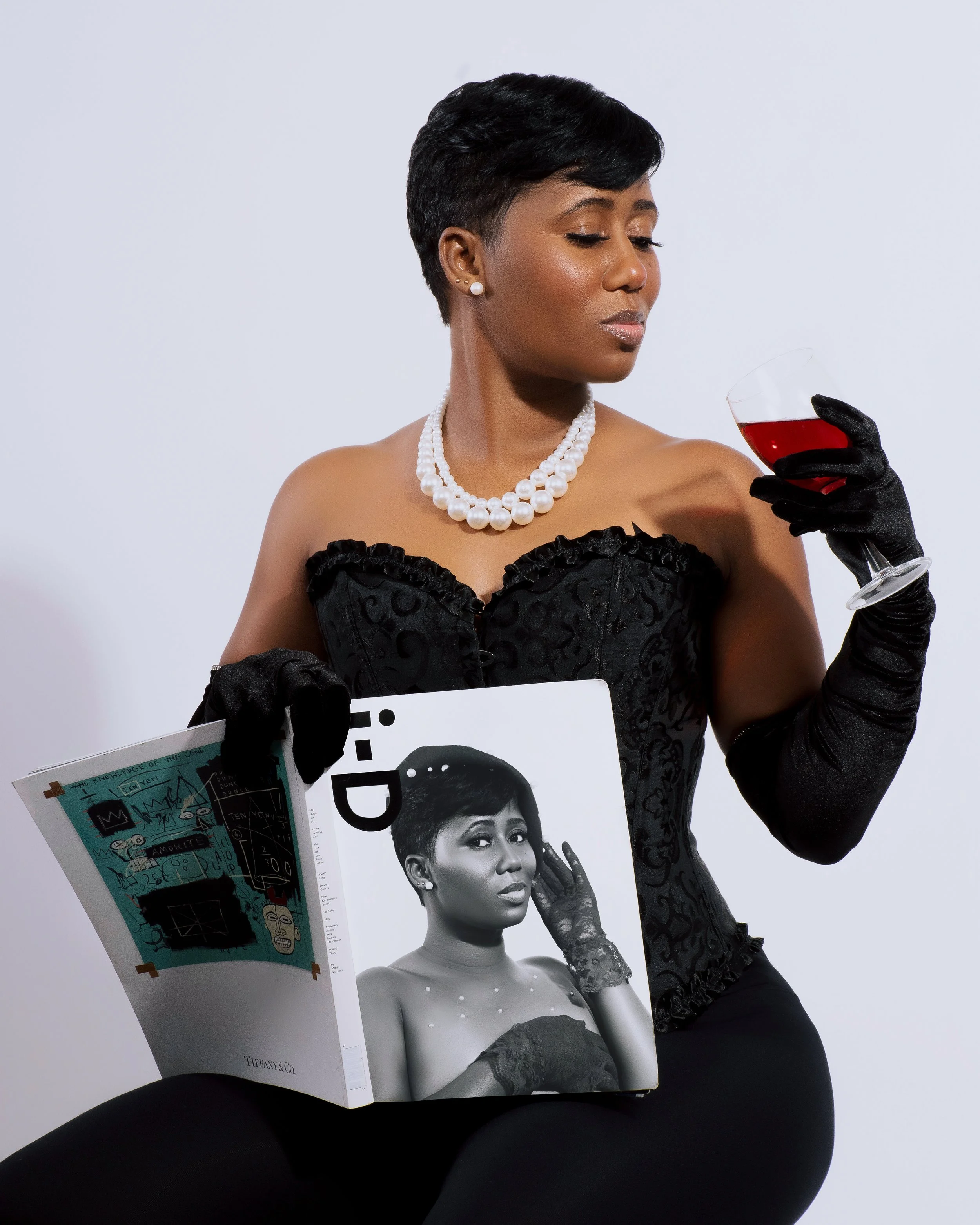 A woman dressed in elegant black attire with pearl jewelry and black gloves, holding a glass of red wine in one hand and a magazine featuring a black and white photo of herself in the other.