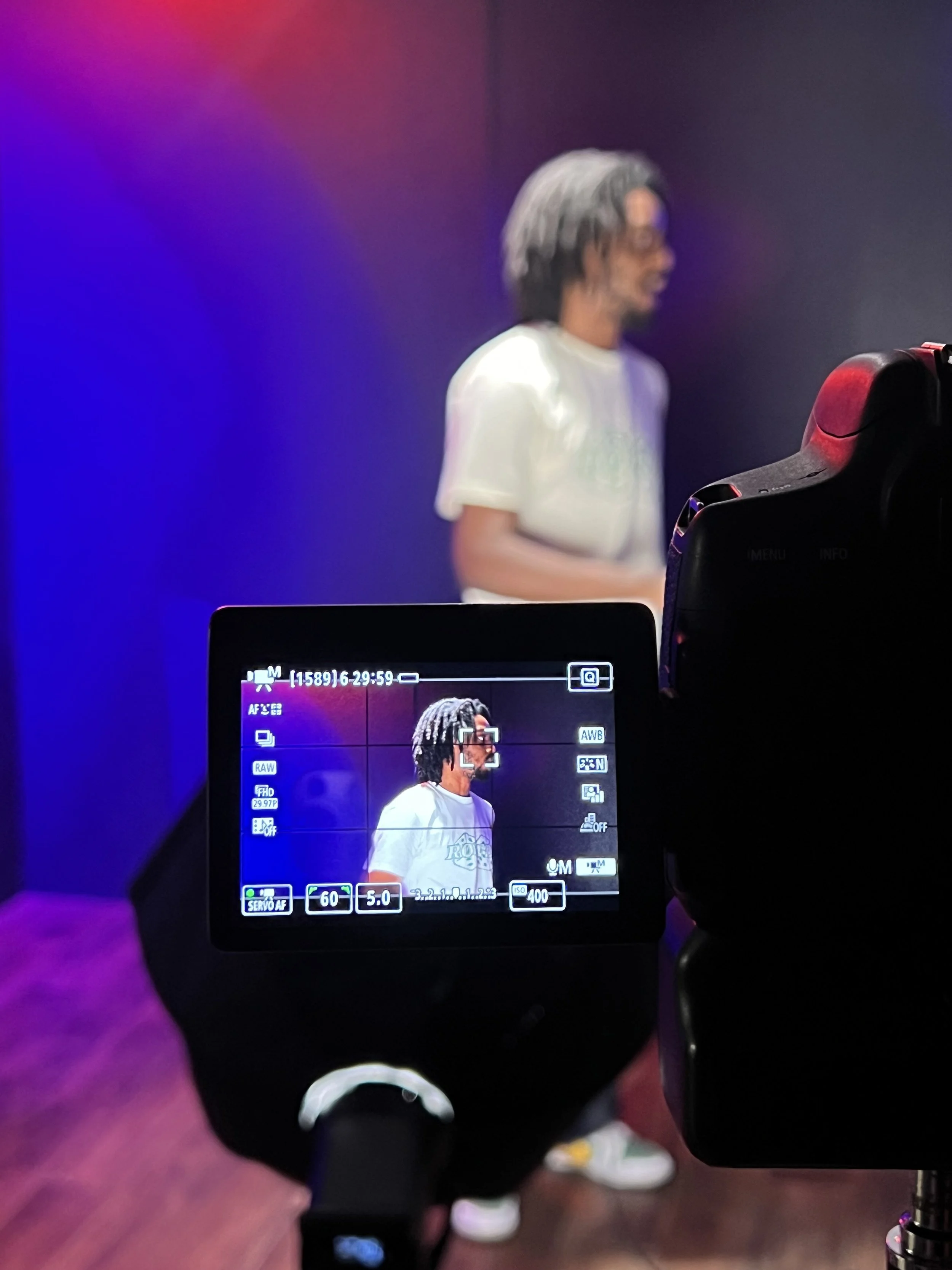A person in a white t-shirt with glasses and dreadlocks is being filmed with a camera on a tripod in a studio with colorful lighting.