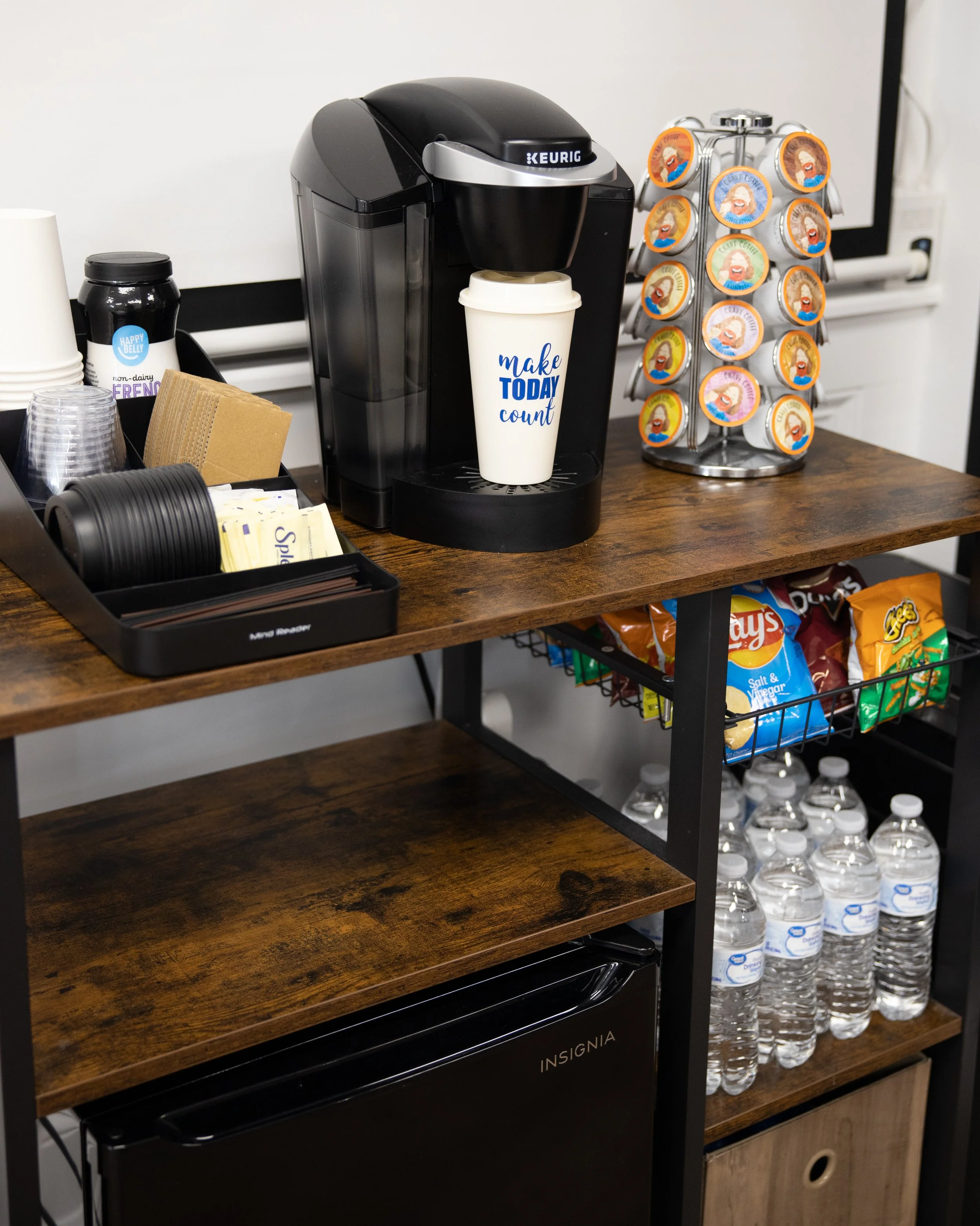 A coffee station with a Keurig machine, a cup with the text 'Make TODAY count,' a container of assorted coffee pods, a tray of disposable cups, napkins, and sugar packets, a small rack with flavored creamer bottles, snack bags, and water bottles underneath.