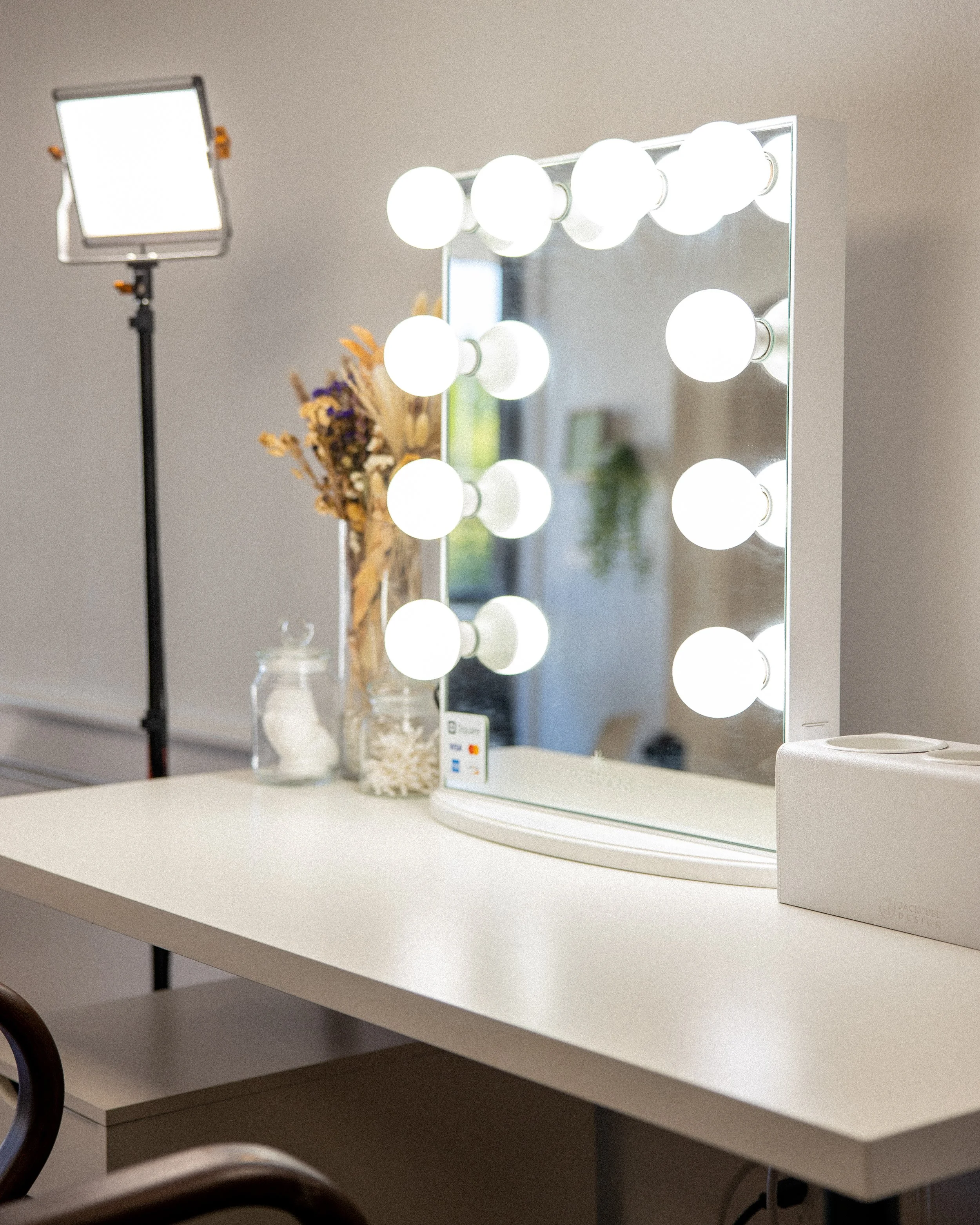 A mirror with multiple round lights around the frame, placed on a white table with dried flowers and jars, in front of a window. Professional lighting equipment is visible in the background.