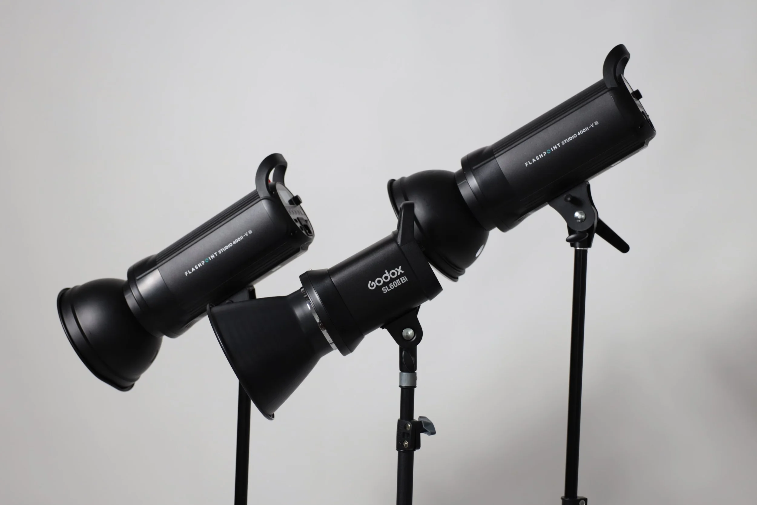 Two black professional studio lights mounted on adjustable stands against a plain gray background.