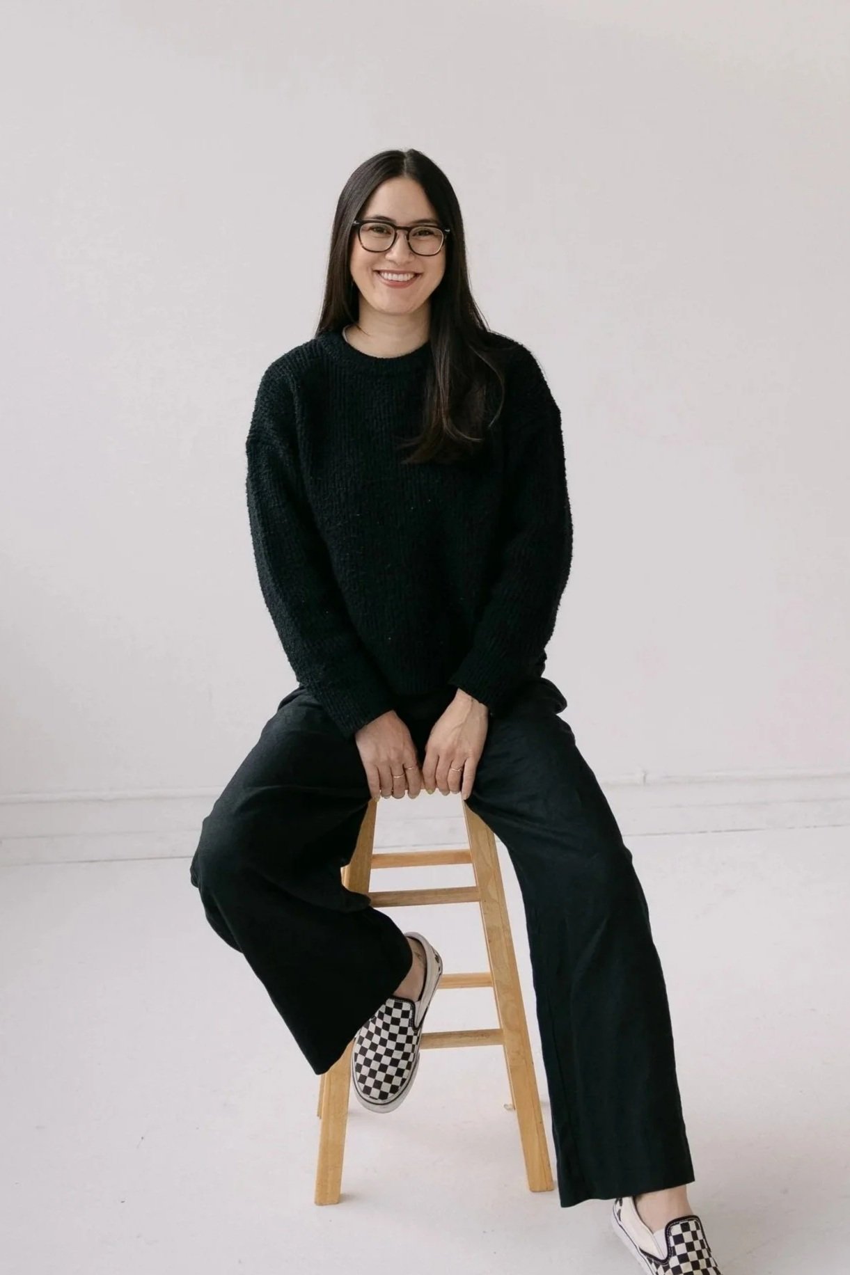 BIPOC female therapist sitting on a wooden stool, wearing glasses, a black sweater, black pants, and checkered slip-on shoes, smiling against a white background.