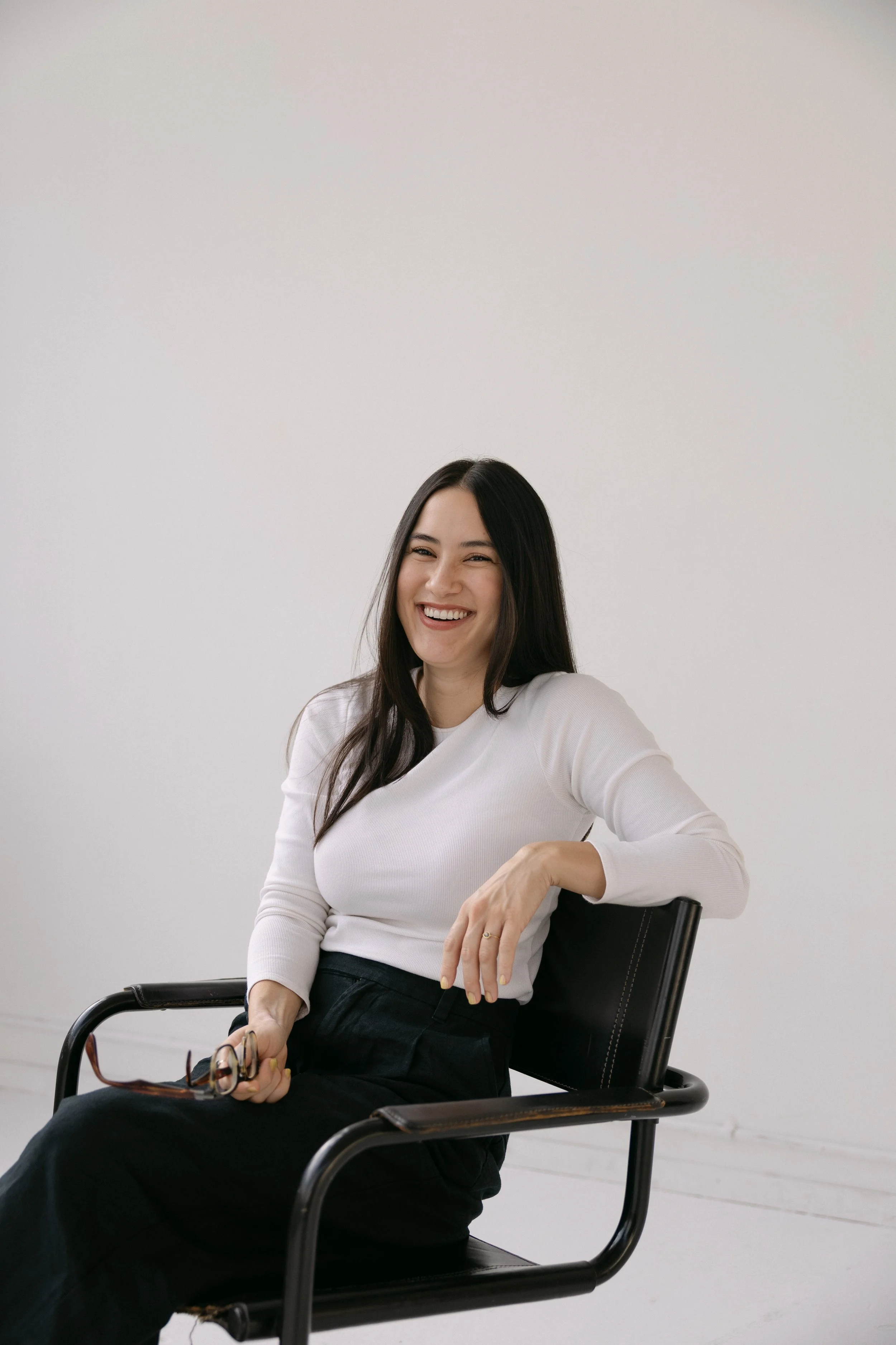 A woman sitting in a black chair, smiling, with long dark hair, wearing a white long-sleeve top and black pants, holding glasses in her right hand, in front of a plain white background.