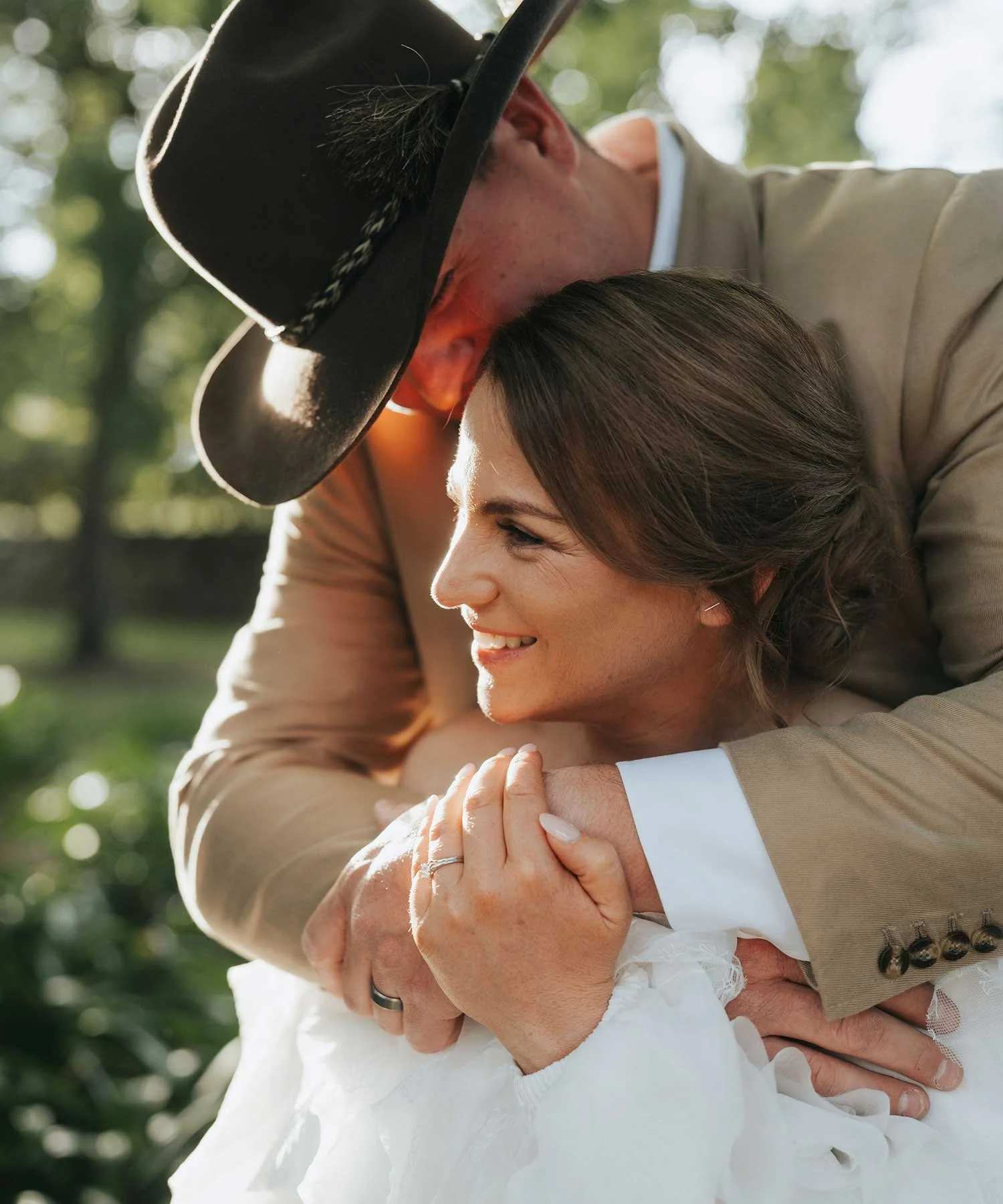 A happy couple sharing a tender embrace outdoors in a sunlit setting, with the man wearing a cowboy hat and the woman smiling.