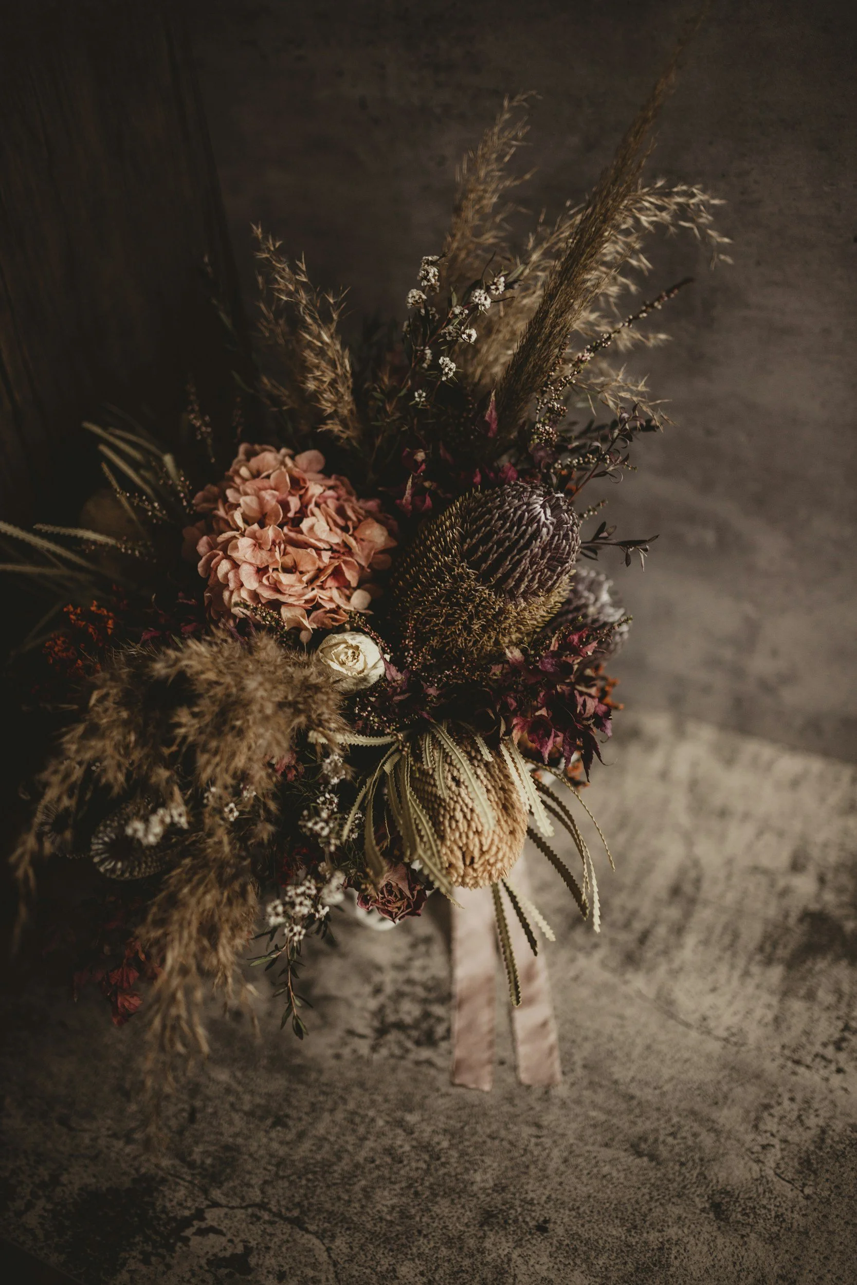 A dried flower bouquet with pink, purple, and beige flowers, along with pampas grass, in front of a dark wall and concrete floor.
