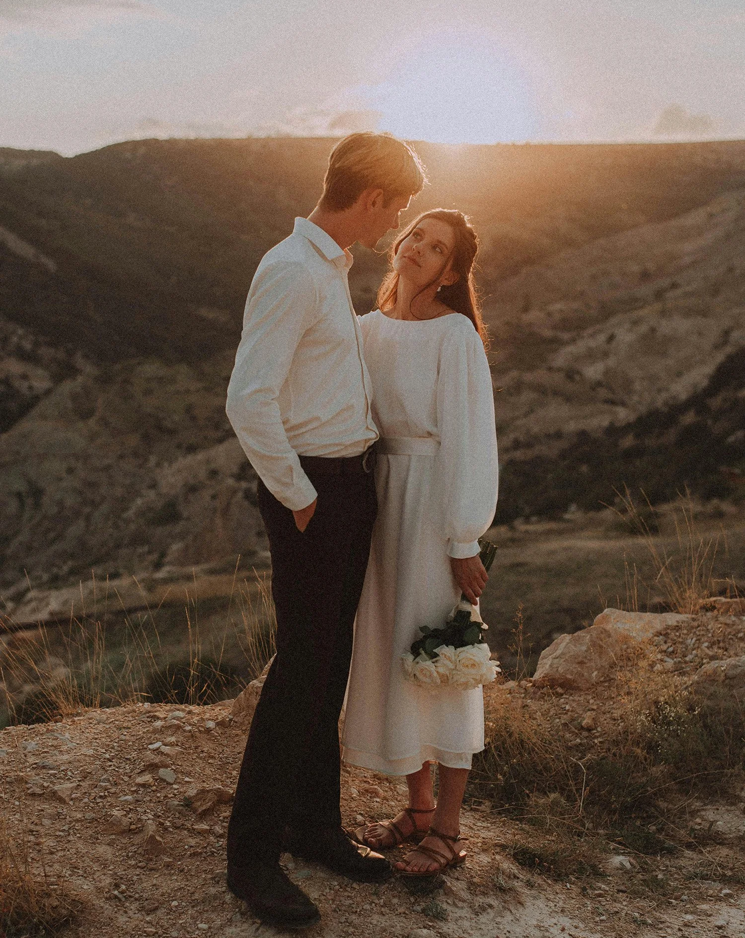 A man and woman stand close together on a rocky hill at sunset, gazing into each other's eyes, with the woman holding a bouquet of white roses.