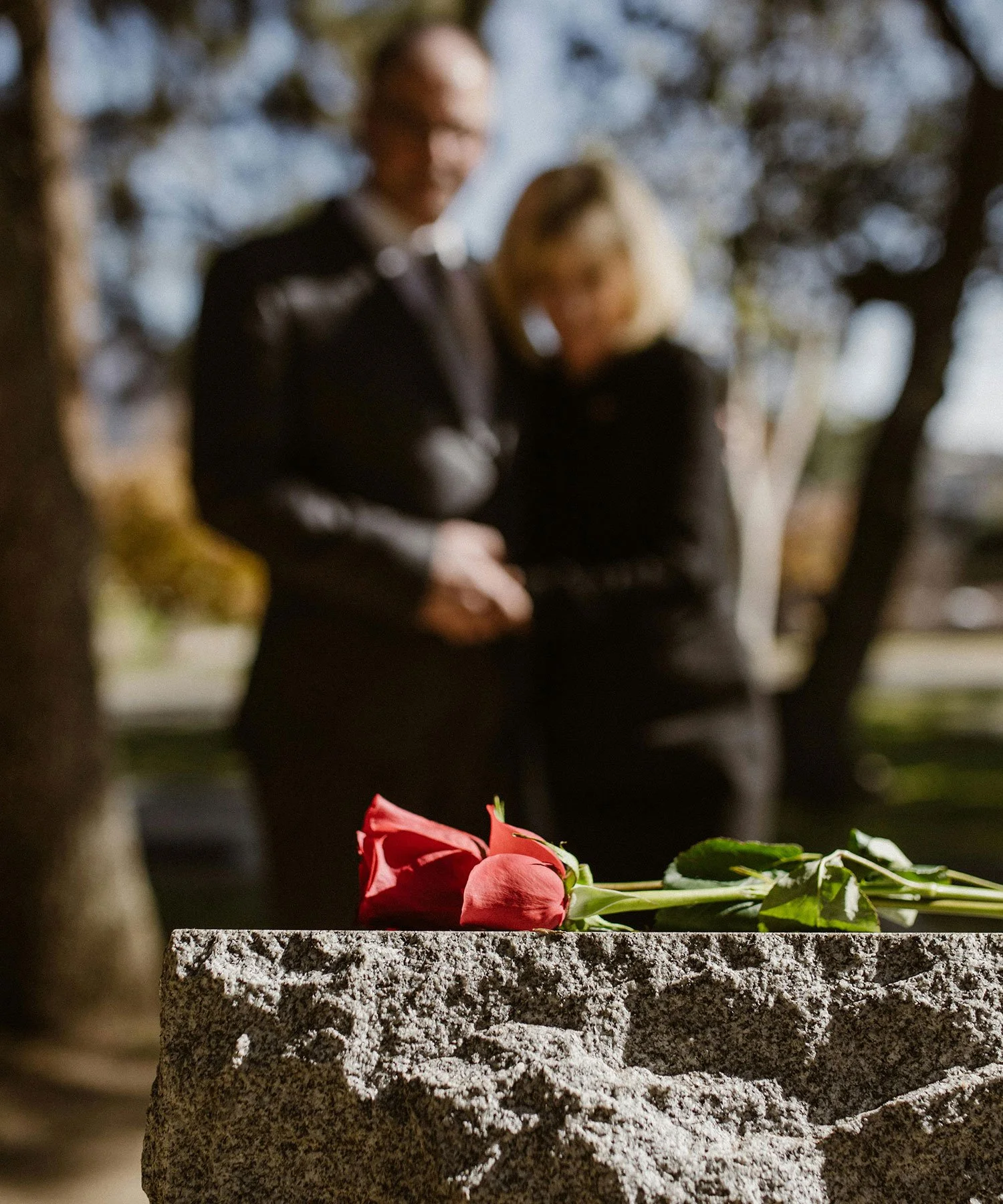 Red long stemmed roses sitting on a grave in cemetery with couple wearing black embracing in the background.