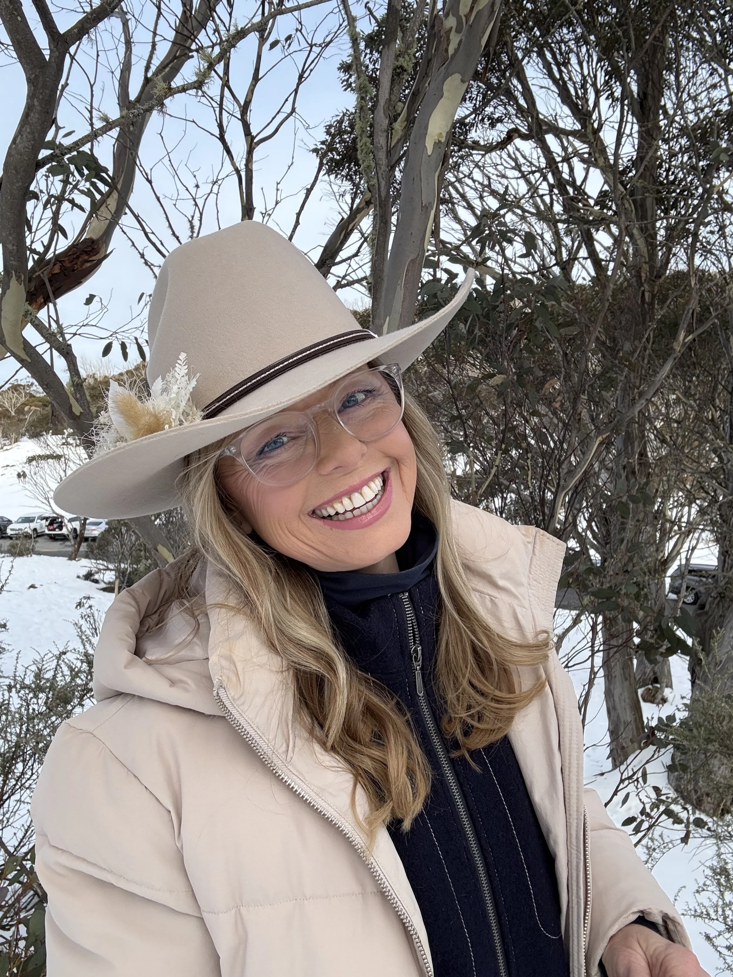 Woman smiling outdoors in winter, wearing a beige coat, a large beige hat with feather and floral embellishments, glasses, and a black fleece jacket, standing in front of leafless trees and snow.