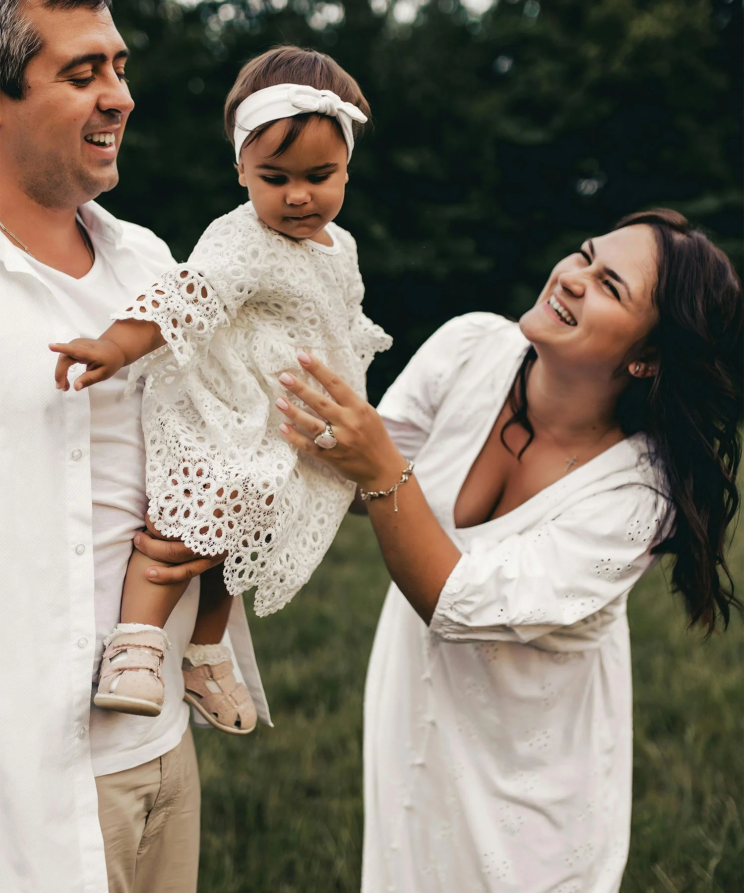 A family of three outdoors, with a man holding a young girl dressed in white with a headband, while a woman with dark hair in a white dress looks at her and smiles.
