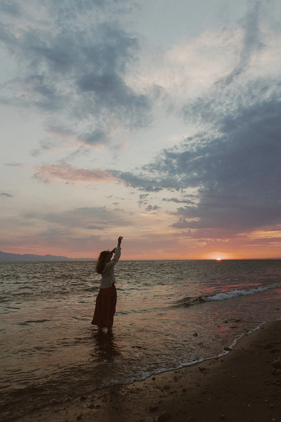 A woman standing in shallow ocean water at sunset, reaching up towards the sky, with a partly cloudy sky above and mountains in the distance.