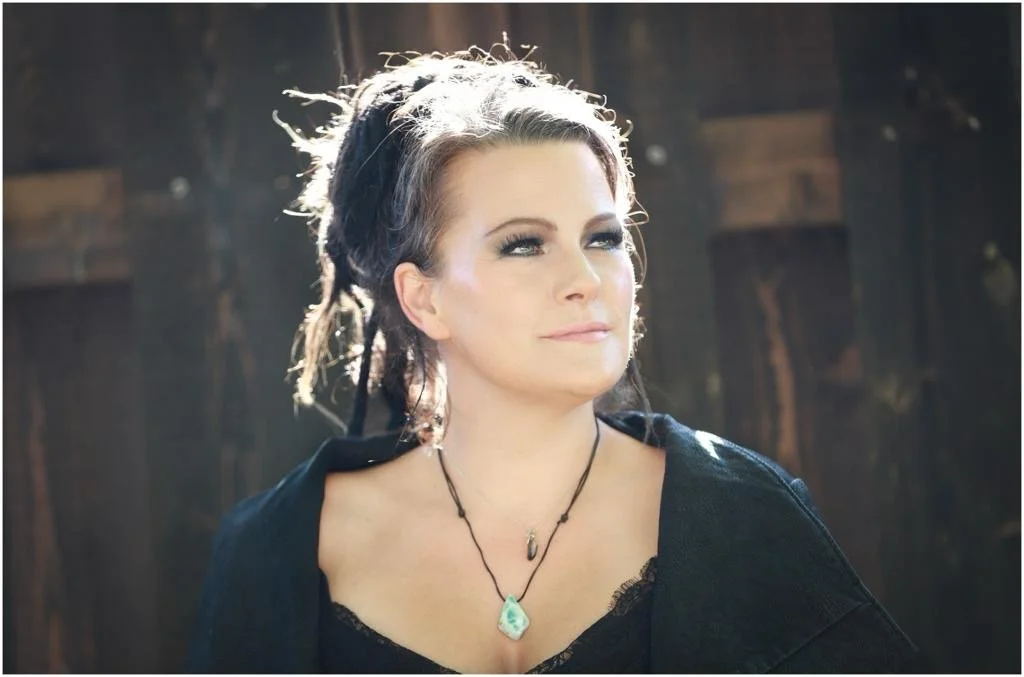 A woman with dark hair styled back, wearing makeup, black sleeveless top, and jewelry, smiling indoors in a casual setting.