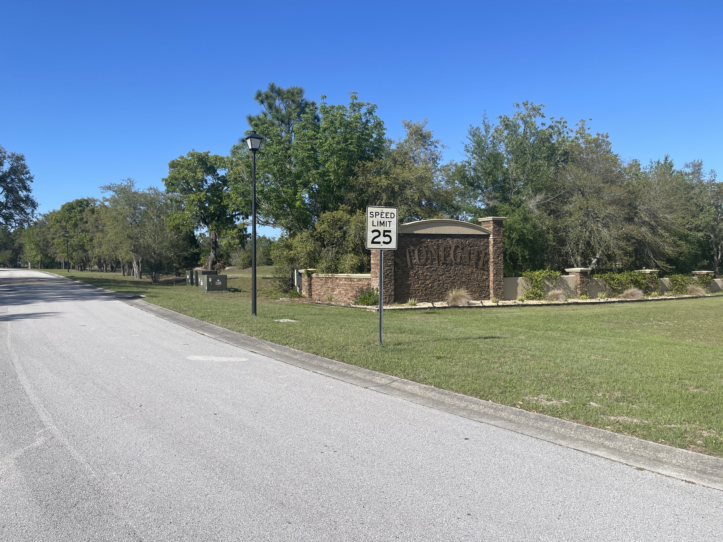 Street view of the entrance to Stonegate neighborhood with a brick sign, a speed limit sign of 25 mph, and trees in the background under a clear blue sky.