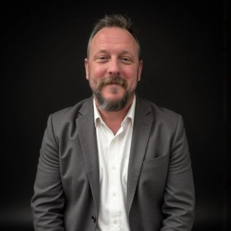 A smiling man with a beard and short hair, wearing a gray suit jacket and white shirt, standing against a black background.