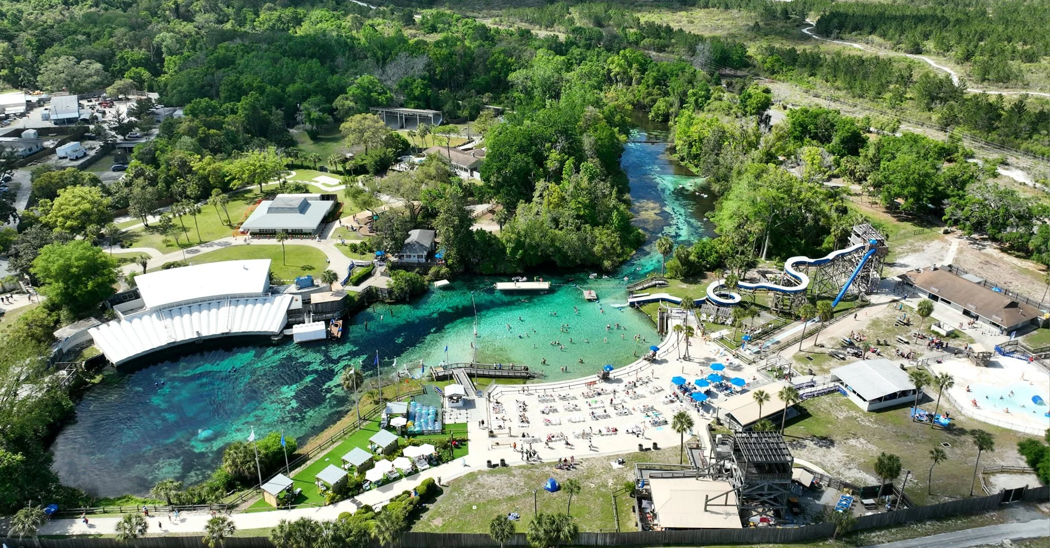 Aerial view of a water park with a large river-like pool, a water slide, and surrounding greenery, recreational buildings, and amenities.