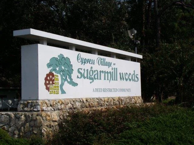 Sign for Cypress Village of Sugarmill Woods on a stone base with a tree and colorful gear logo, indicating a deed-restricted community.