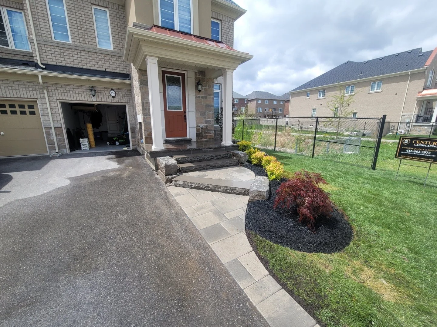 Front porch of a two-story house with red door, stone steps, and a small garden with bushes. The house has a brick exterior and a garage with an open door showing tools and equipment inside. The driveway is asphalt, and there is a black fence with a sign in the yard.