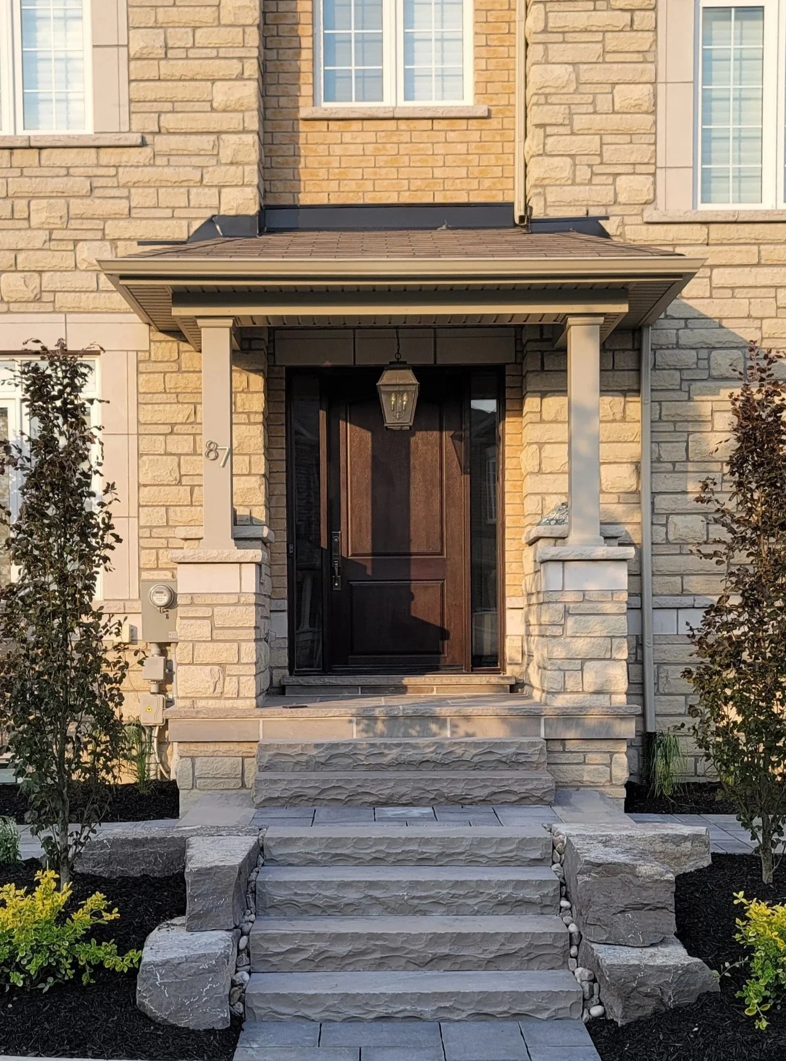 Front entrance of a house with stone steps leading to a dark wooden door, flanked by columns, with a lantern hanging above the door, and shrubbery on both sides.