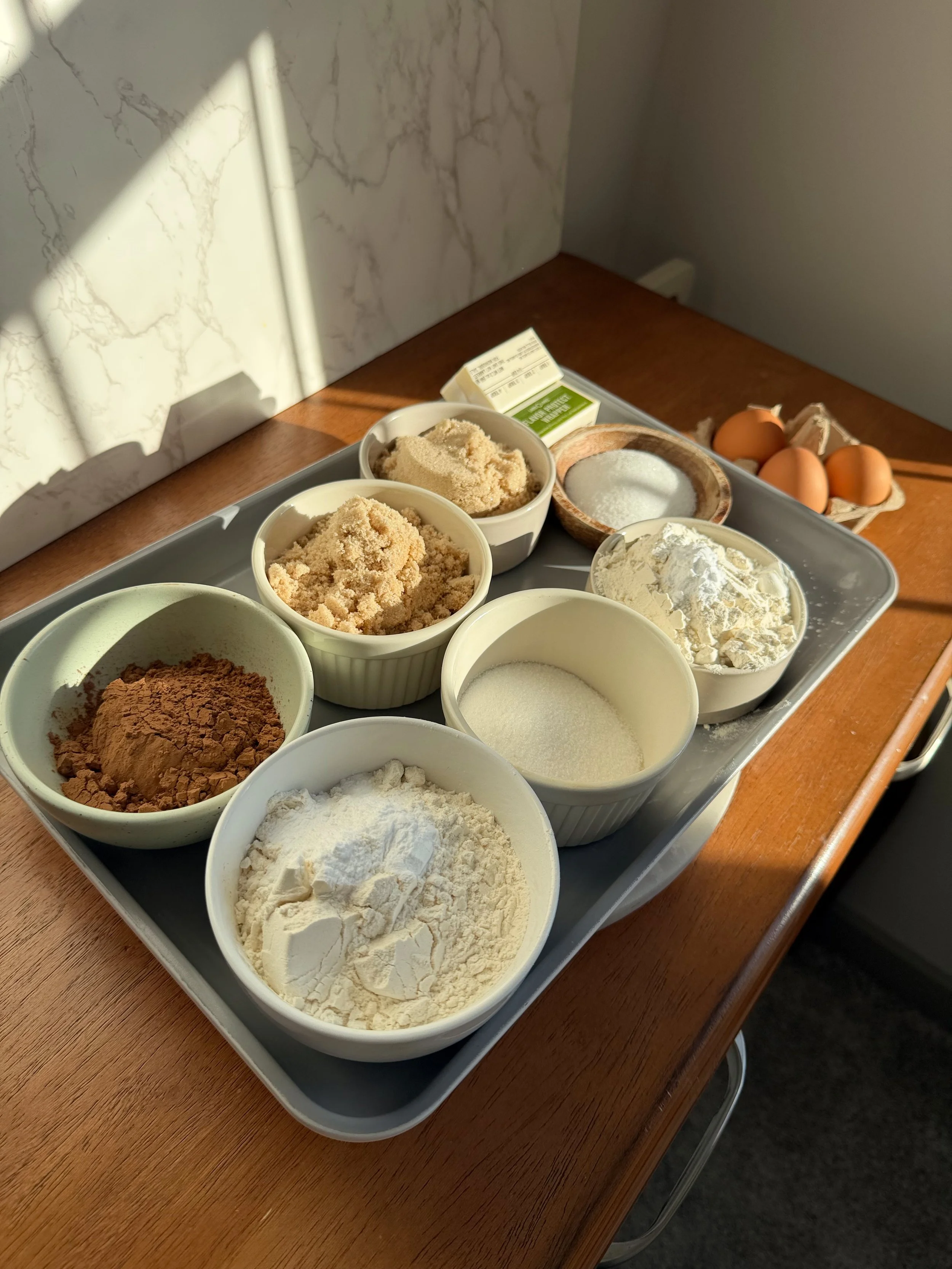 Ingredients for brookie cookies laid out on a counter with butter, sugars, eggs, cocoa, flour, chocolate chips, and brownie batter ready to mix.