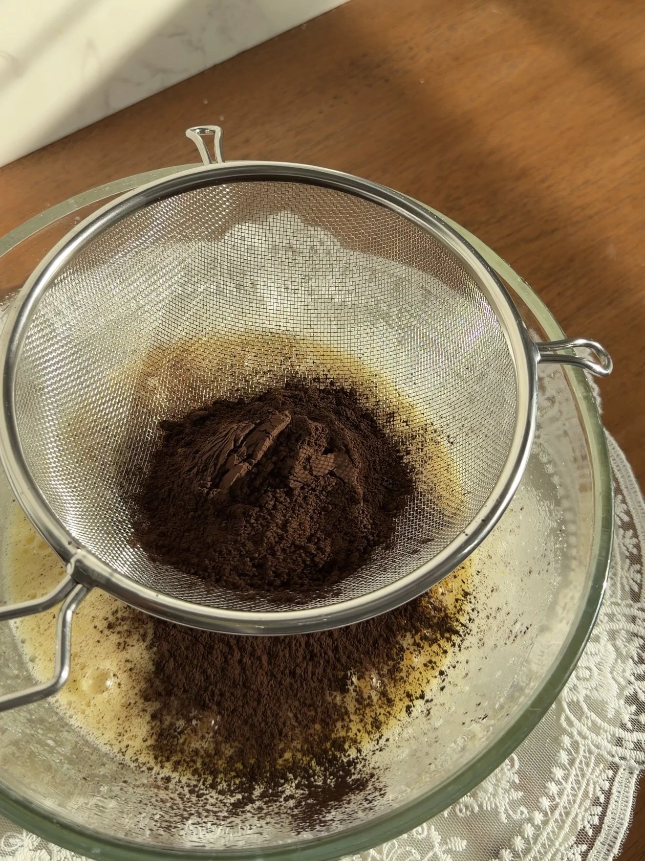 Cocoa powder being sifted into glass mixing bowl on the counter.