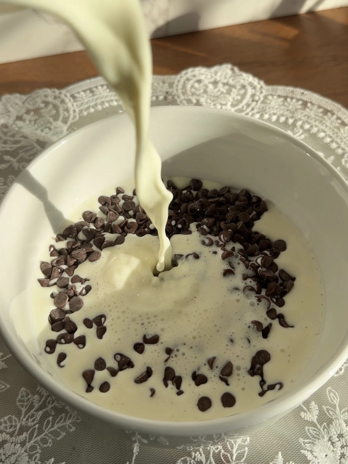 Warm heavy cream being poured over chocolate chips in a small white mixing bowl. 
