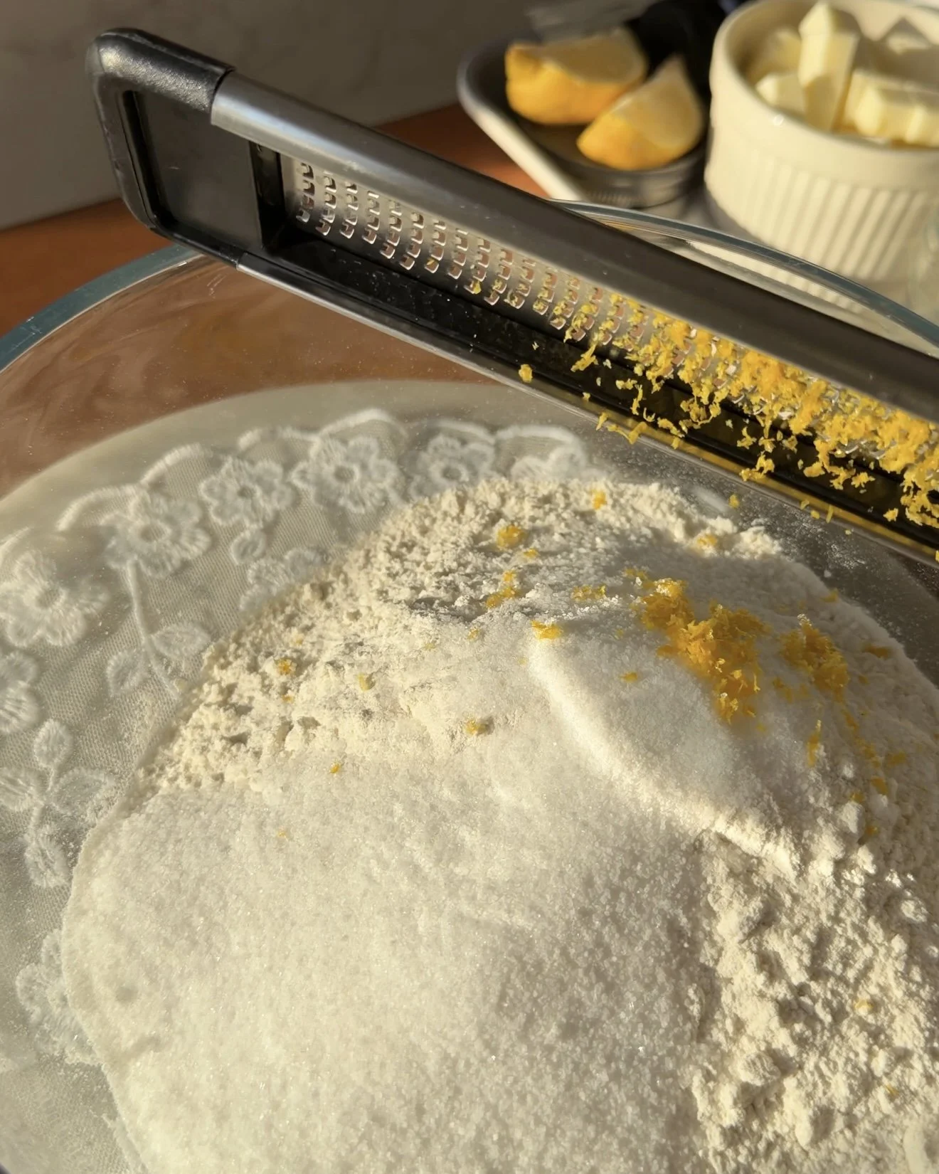 Glass mixing bowl on countertop with dry ingredients , sugar, and lemon zest