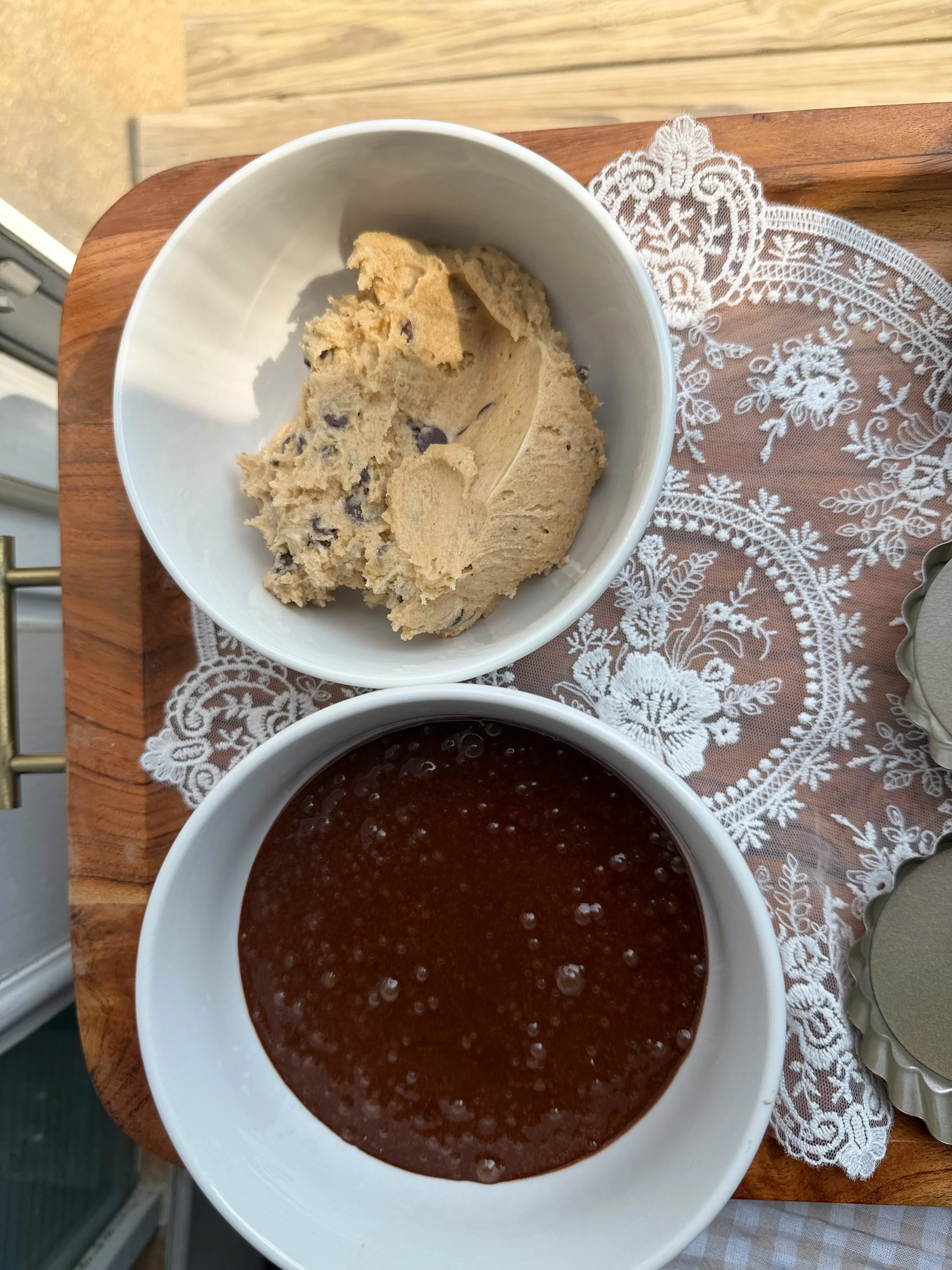 two white bowls with cookie dough and brownie dough on a cutting board. 