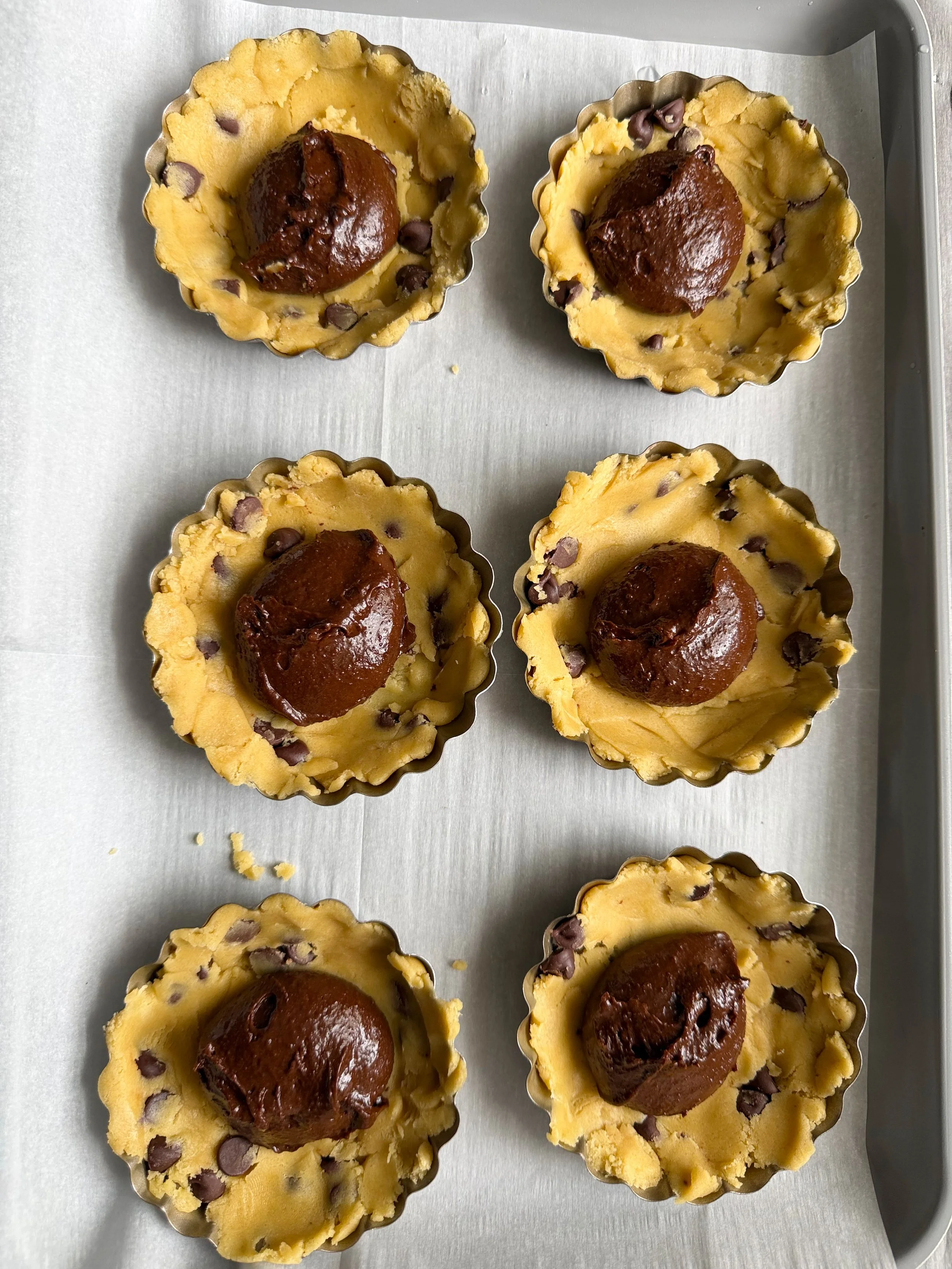 Six tart pans on the counter with brownie scoop on top of even cookie dough