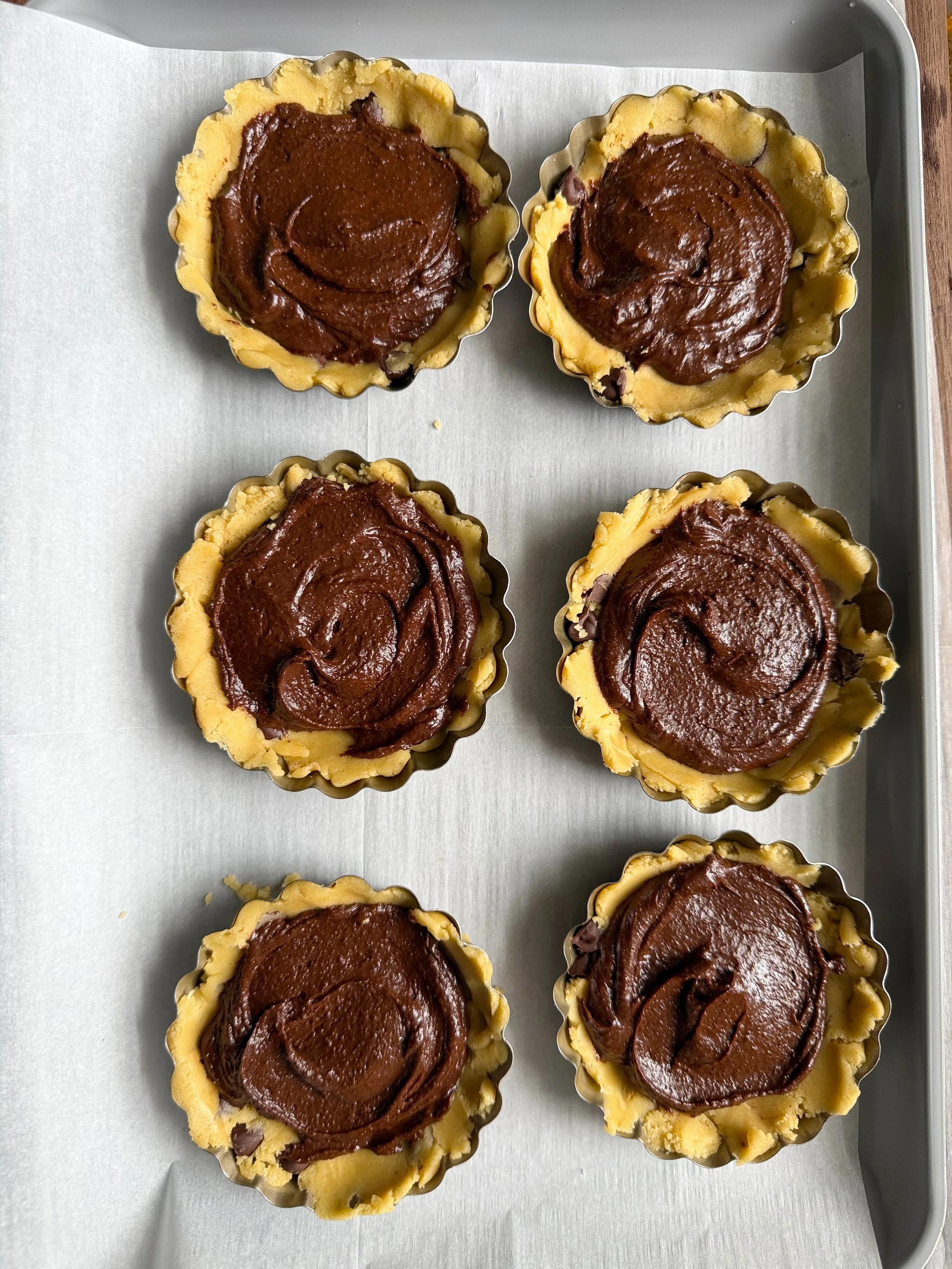 Six tart pans on the counter with brownie evenly spread over cookie dough. 