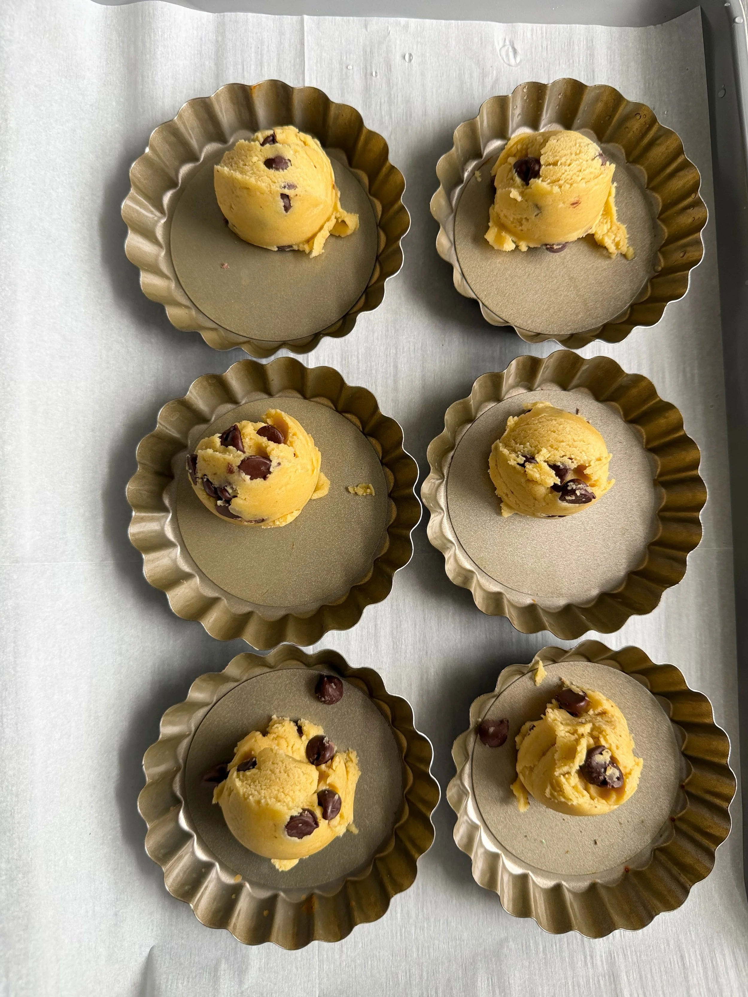 Six tart pans on the counter with one cookie dough scoop in each. 