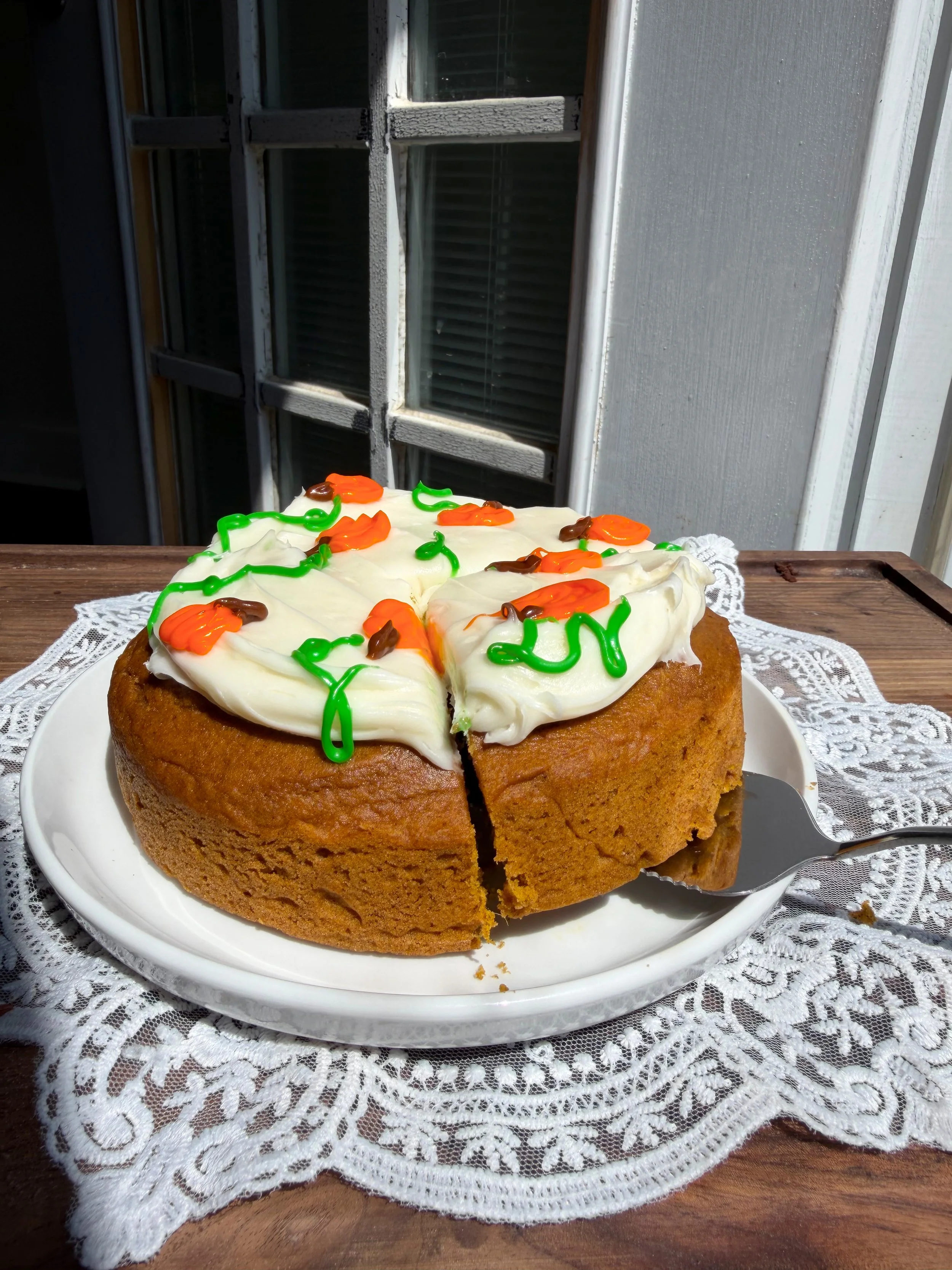 Orange, green, and brown frosting is being piped onto the cream cheese frosted pumpkin cake to create small pumpkins, vines, and stems.