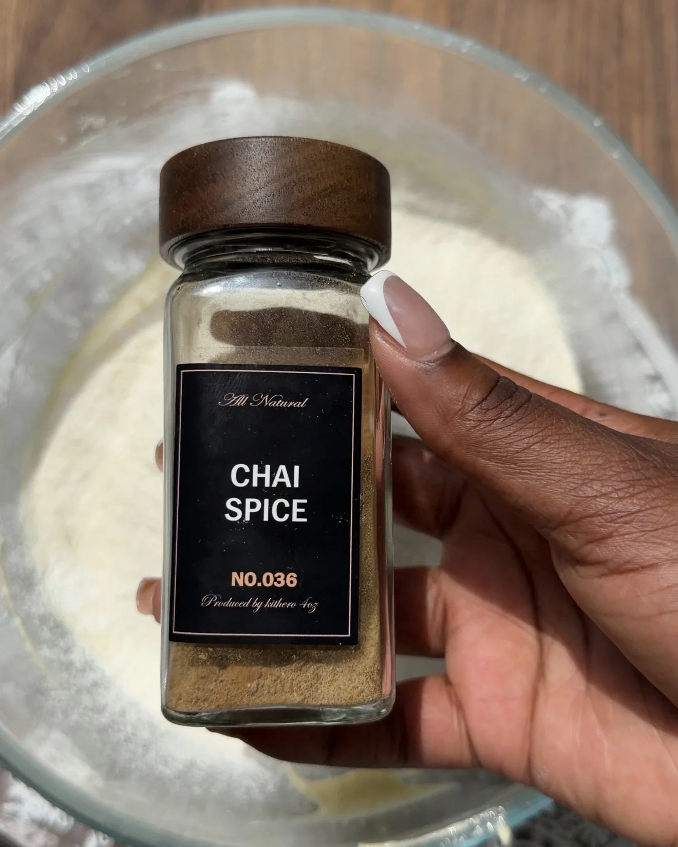 Hand holding chai spice mix container over glass mixing bowl