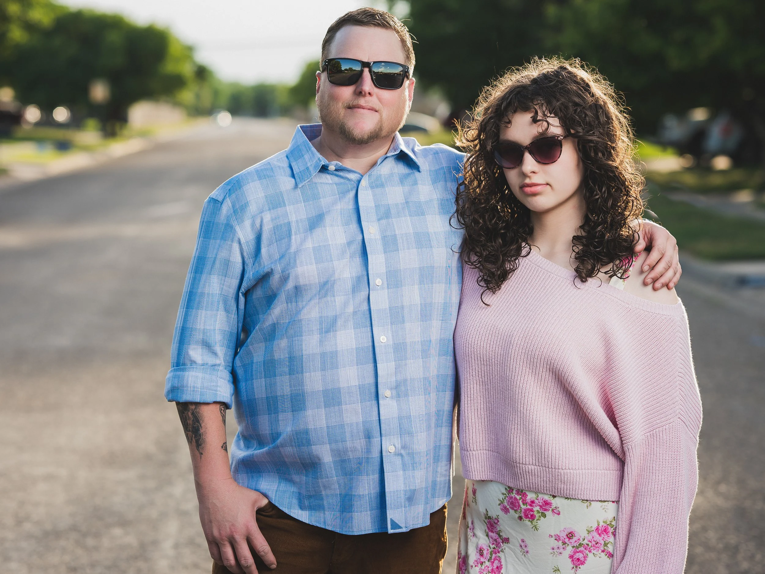 A man and woman stand together outdoors on a sunny day, both wearing sunglasses; the man has a beard, short hair, and a blue checkered shirt with rolled-up sleeves, and the woman has curly dark hair, a pink off-shoulder sweater, and a floral skirt.