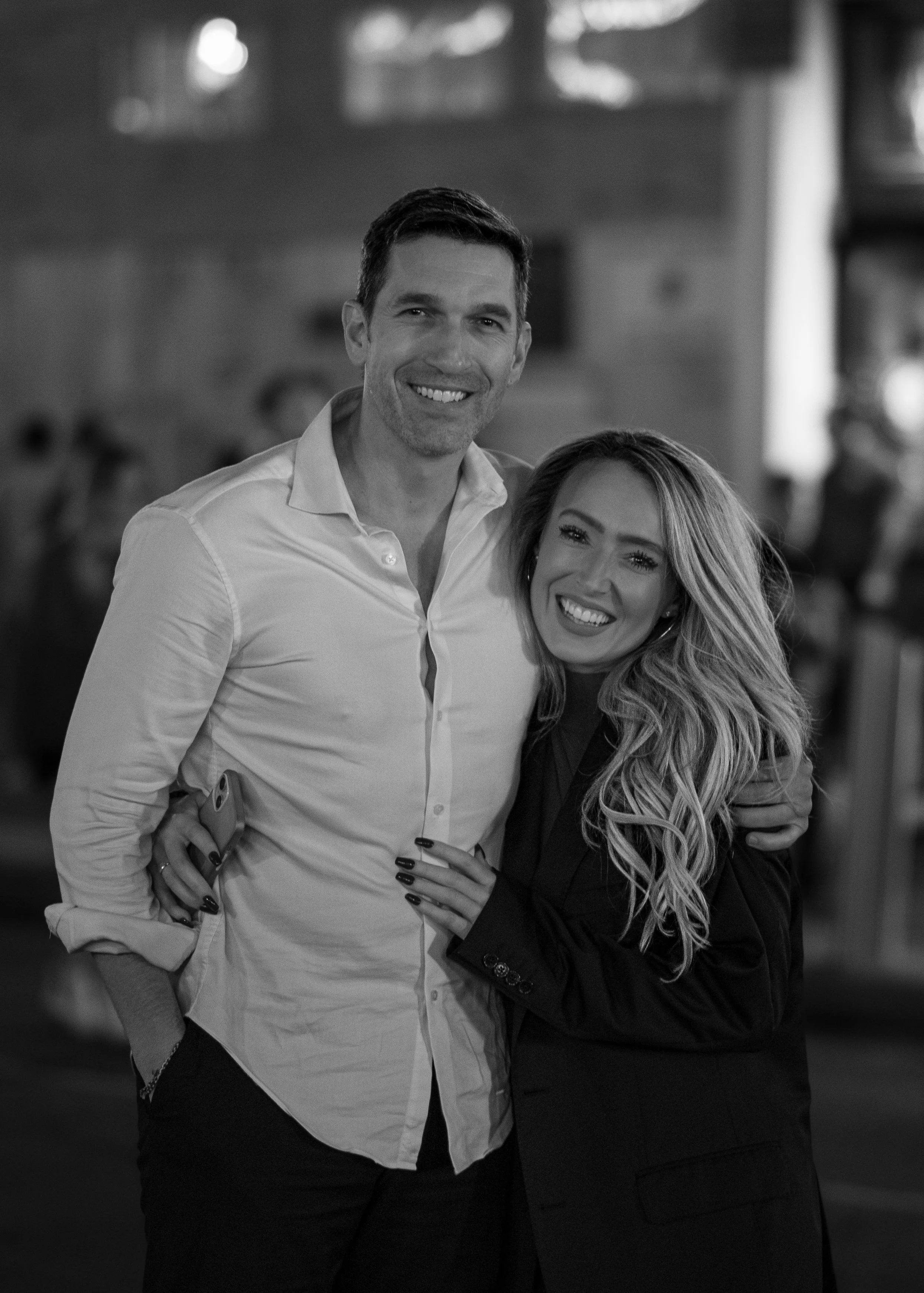 A smiling man and woman pose together indoors, with the man holding a phone in his pocket, both dressed in smart casual wear, the woman with long wavy hair wearing a blazer.