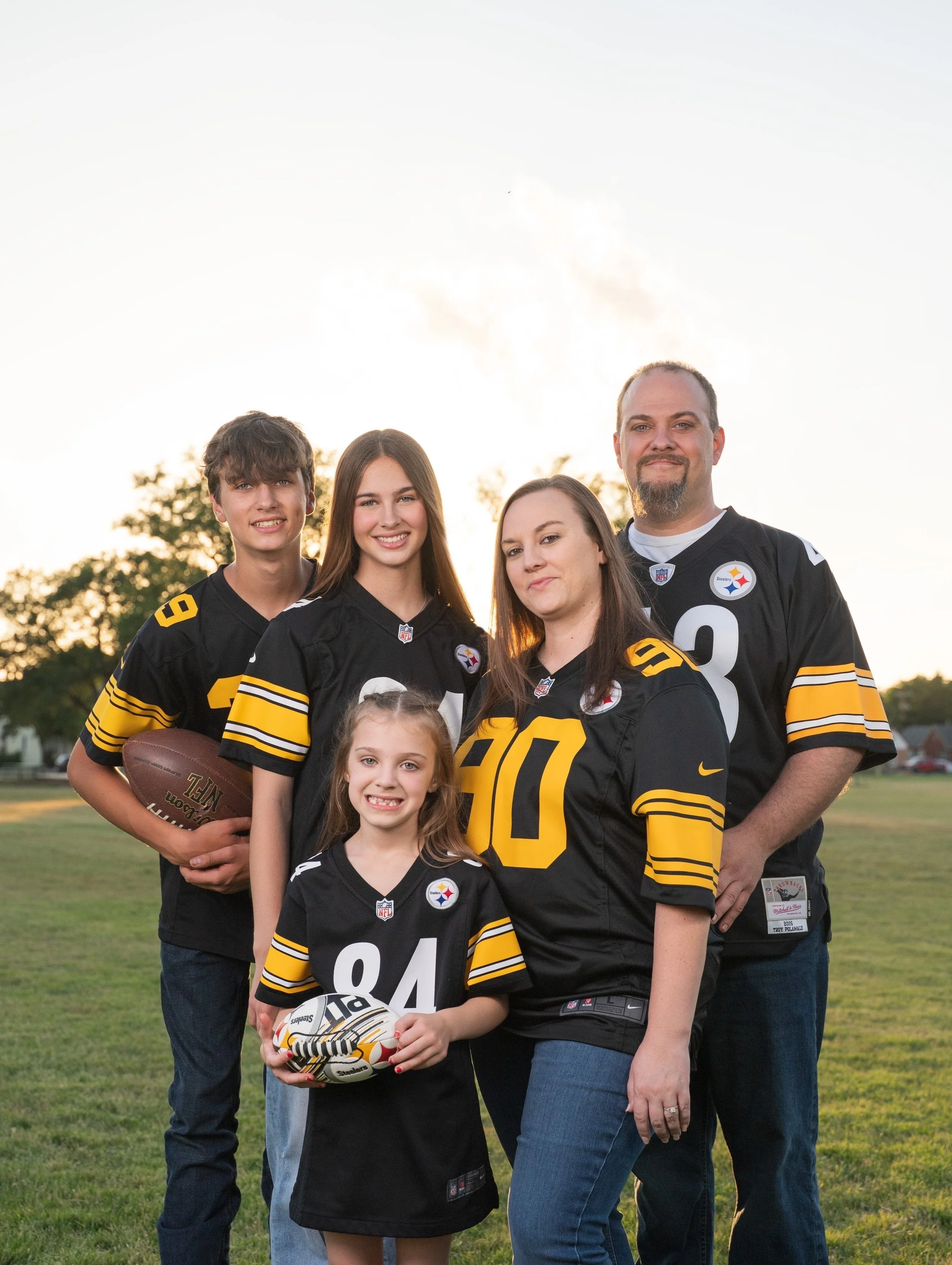 A family of five wearing Pittsburgh Steelers football jerseys posing outdoors on a grassy field during sunset.