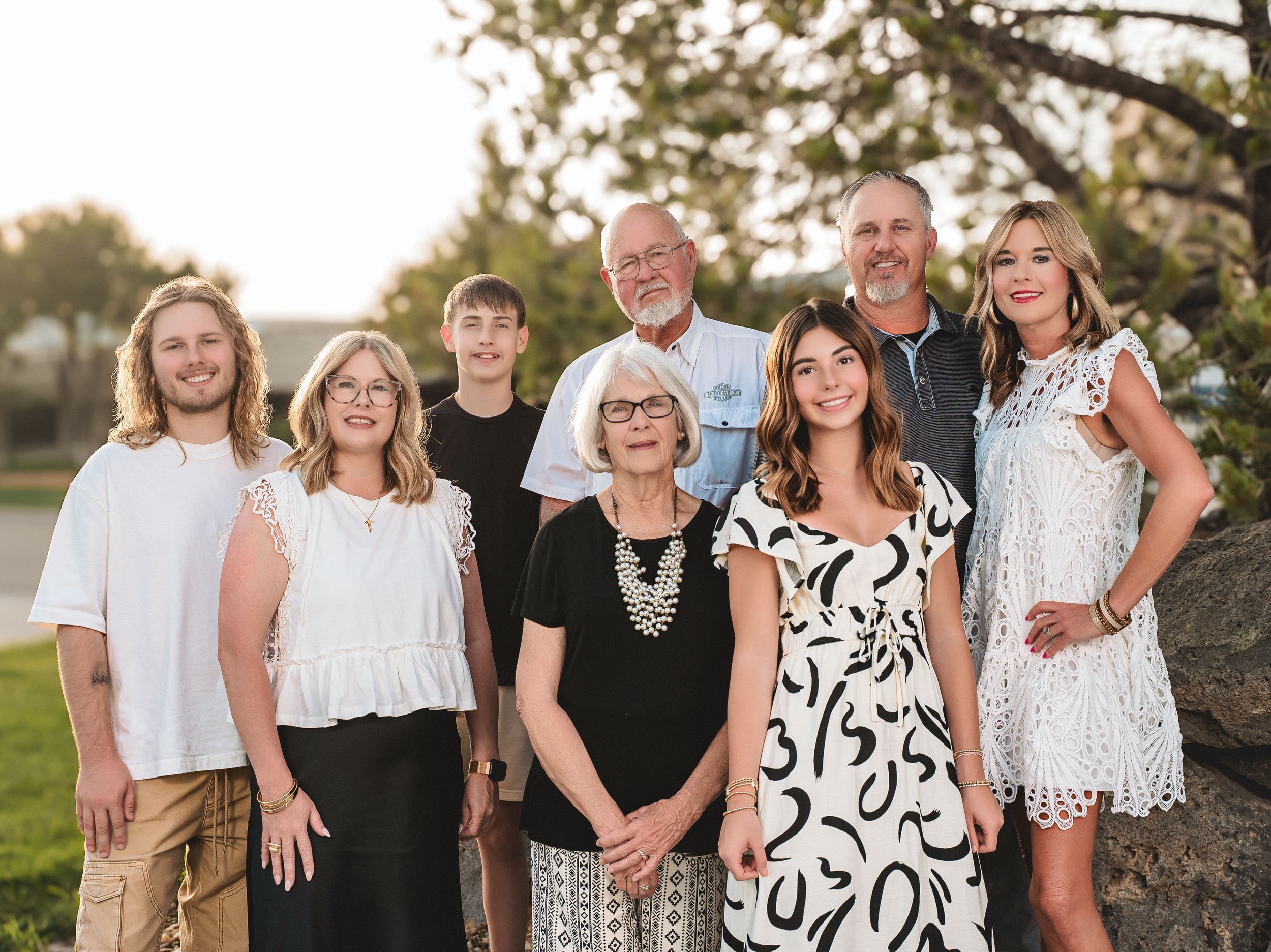 A multi-generational family group portrait outdoors, with nine people smiling and standing together on a grassy area with trees in the background, during sunset.