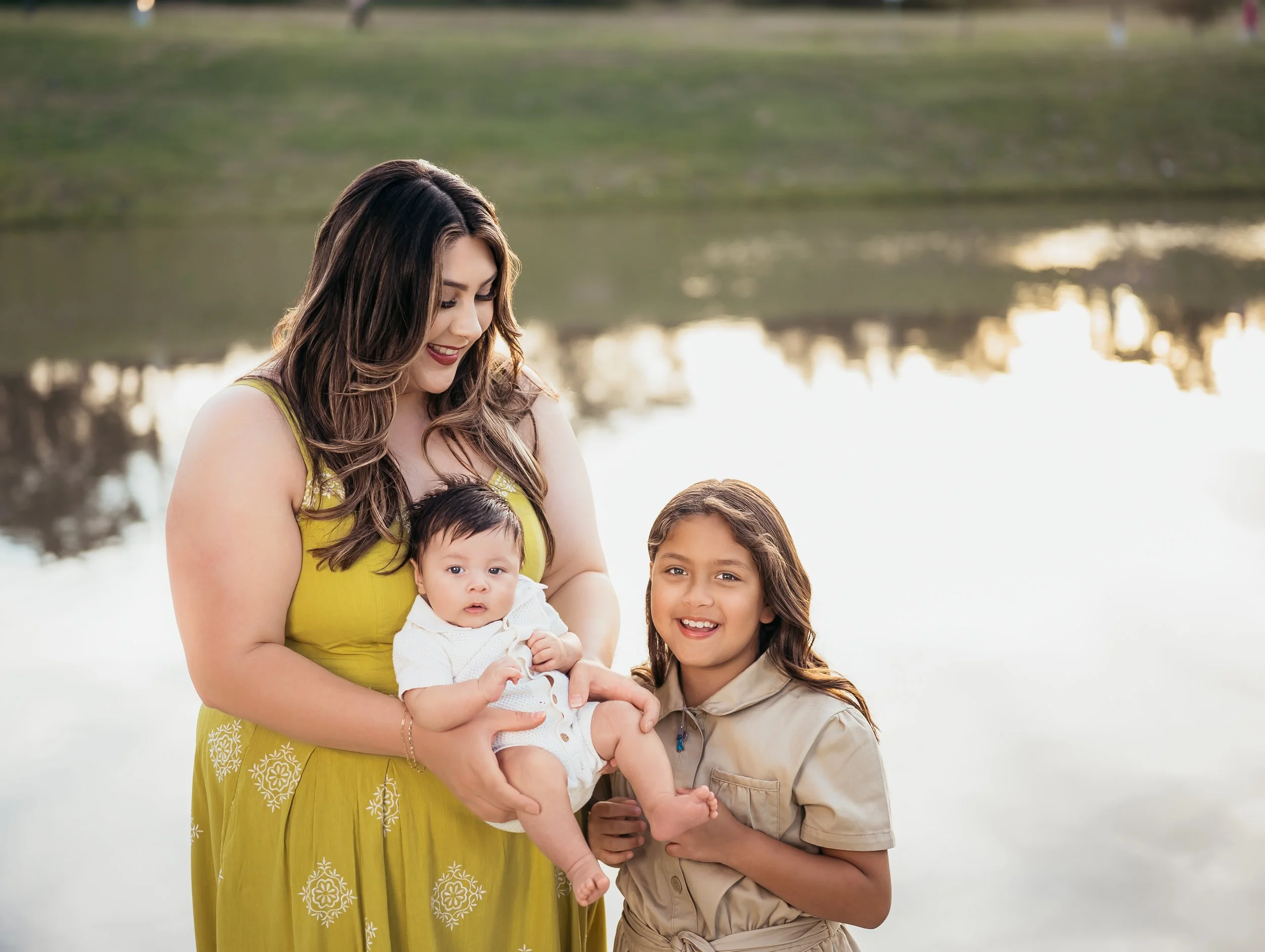 A woman holding a baby and smiling at a young girl by a lake during sunset.
