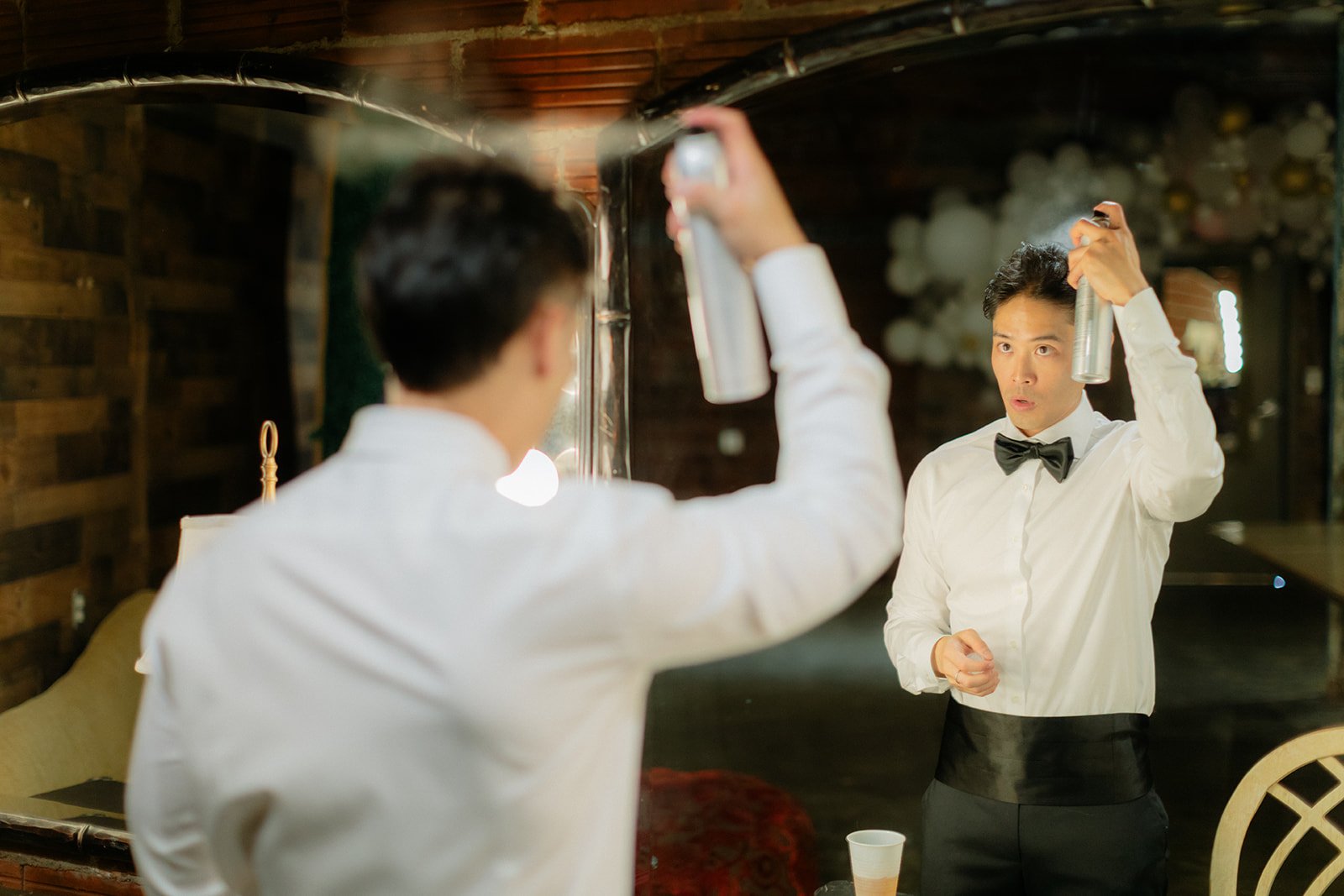 A man in formal attire wearing a white shirt and black bow tie, standing in front of a mirror, sprays hair with a can of hairspray, looking at his reflection in a mirror, in a room with wooden walls and balloons in the background.