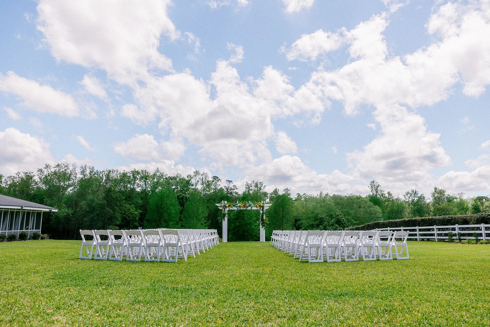 Outdoor wedding setup with white chairs facing a decorated arch on a grassy lawn under a partly cloudy sky, surrounded by trees and a white fence.
