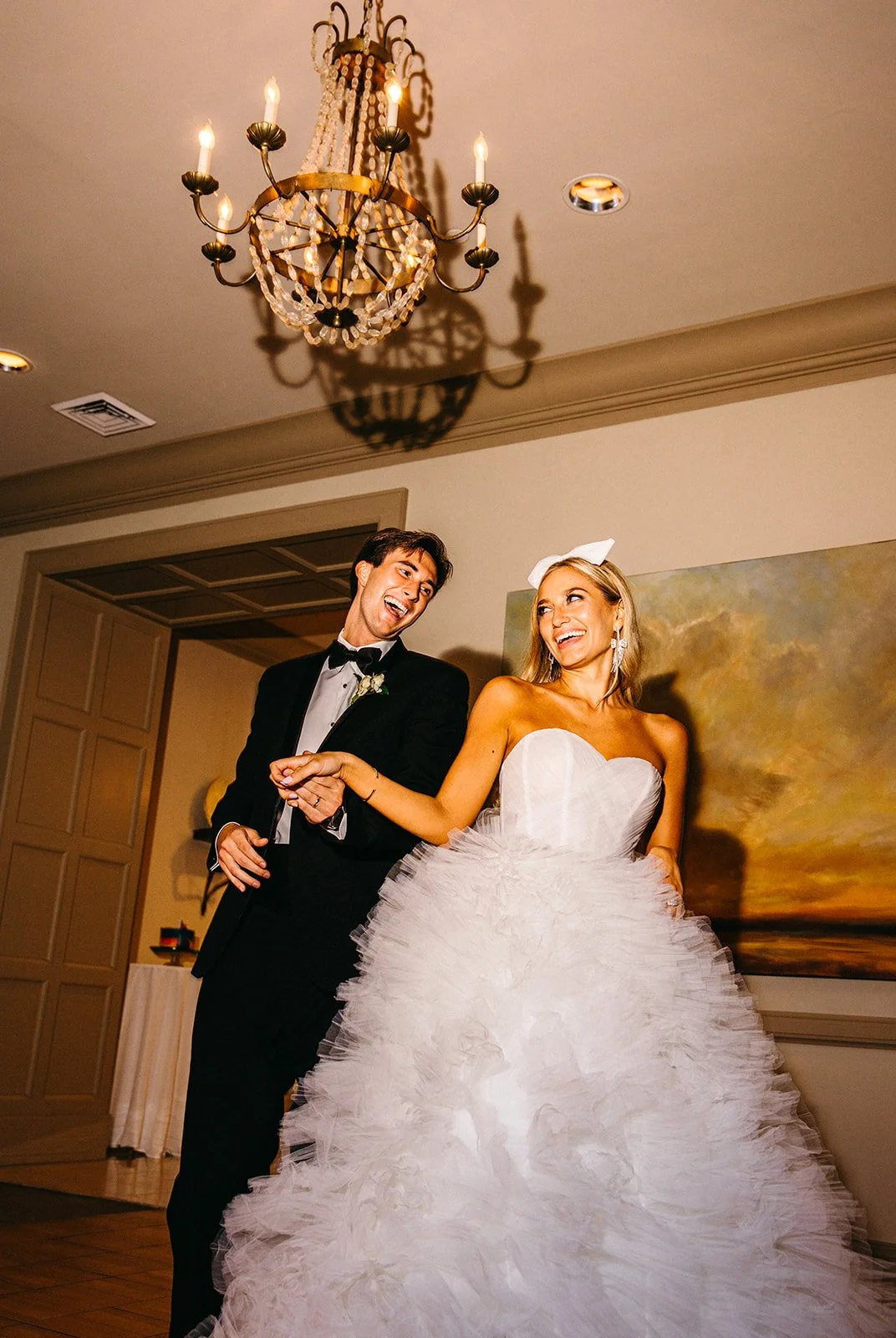 A newlywed couple dancing at their wedding reception. The groom is wearing a black tuxedo, and the bride is wearing a white, strapless wedding gown with a layered, ruffled skirt. They are smiling and holding hands under a chandelier.