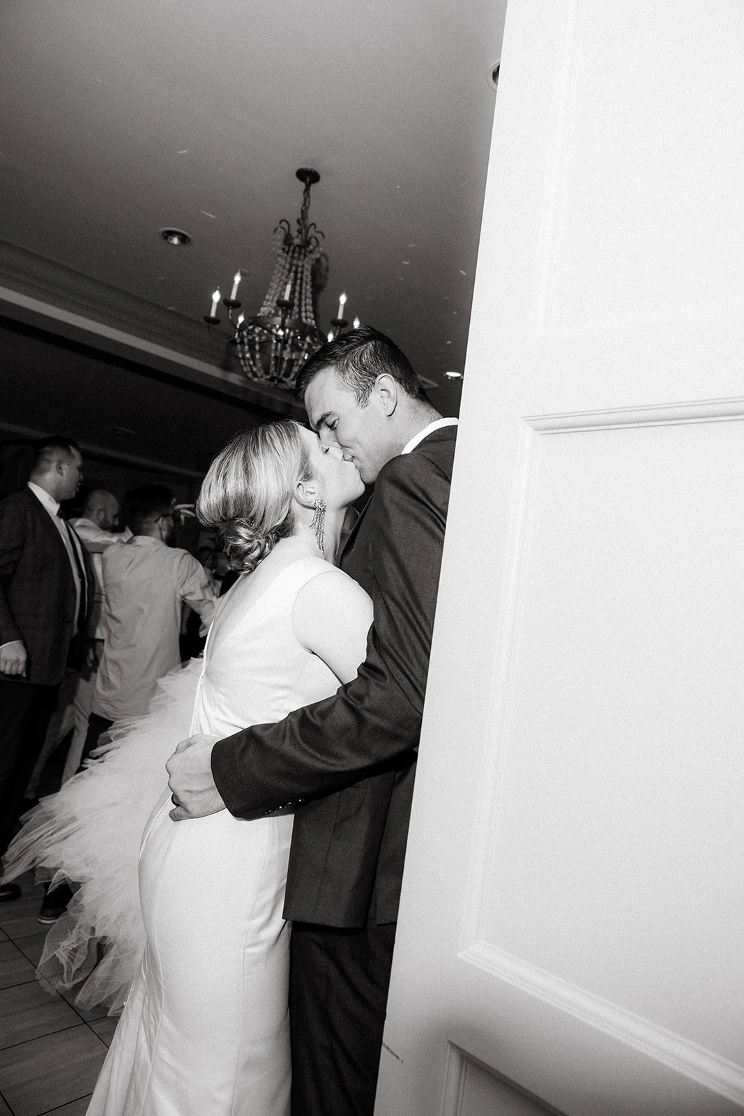 A bride and groom kiss at their wedding reception, standing close together with the bride in a white wedding dress and the groom in a dark suit, in a room with a chandelier and other guests in the background.
