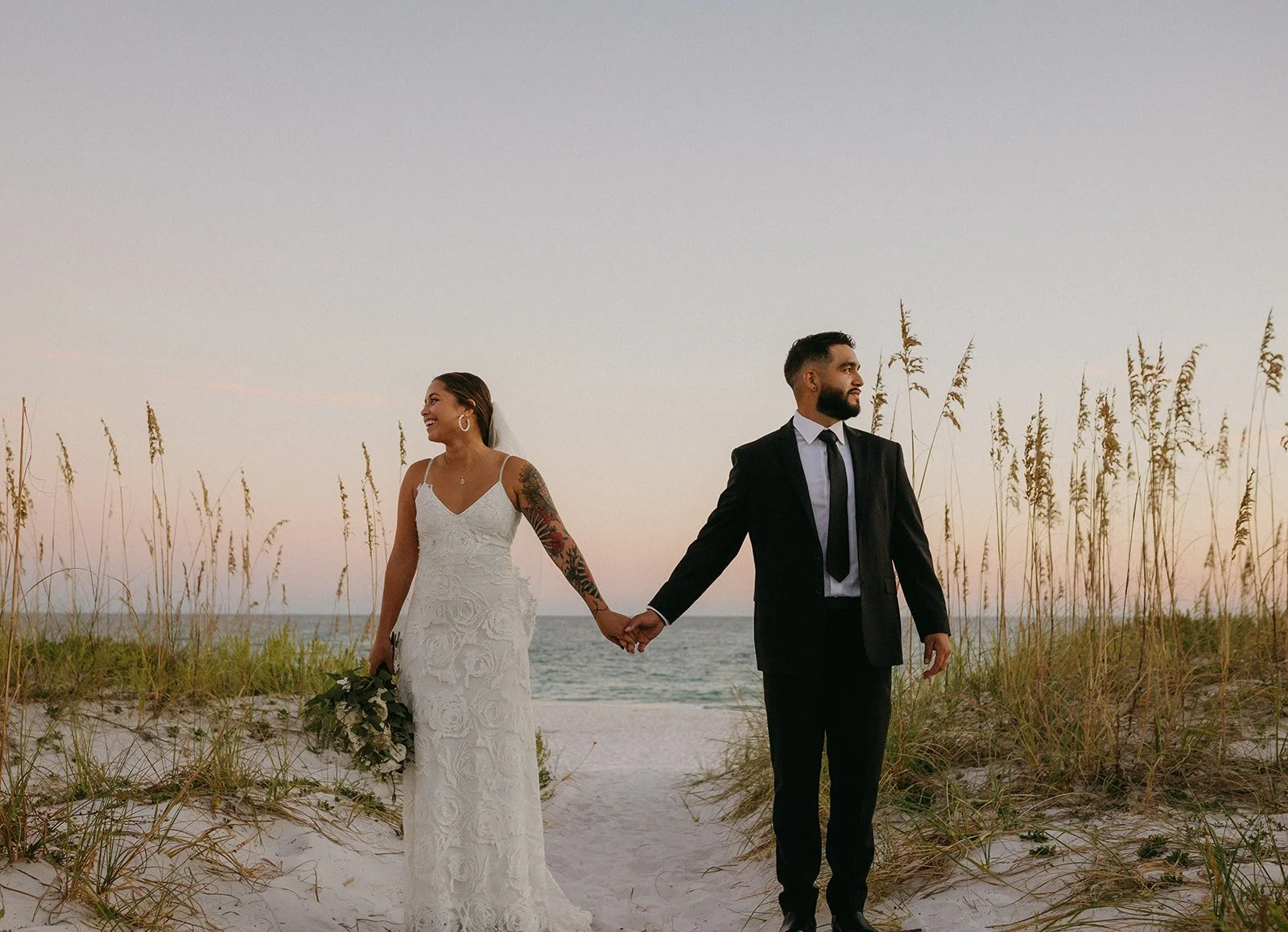A couple in wedding attire holding hands on the beach during sunset, with tall grass and ocean in the background.